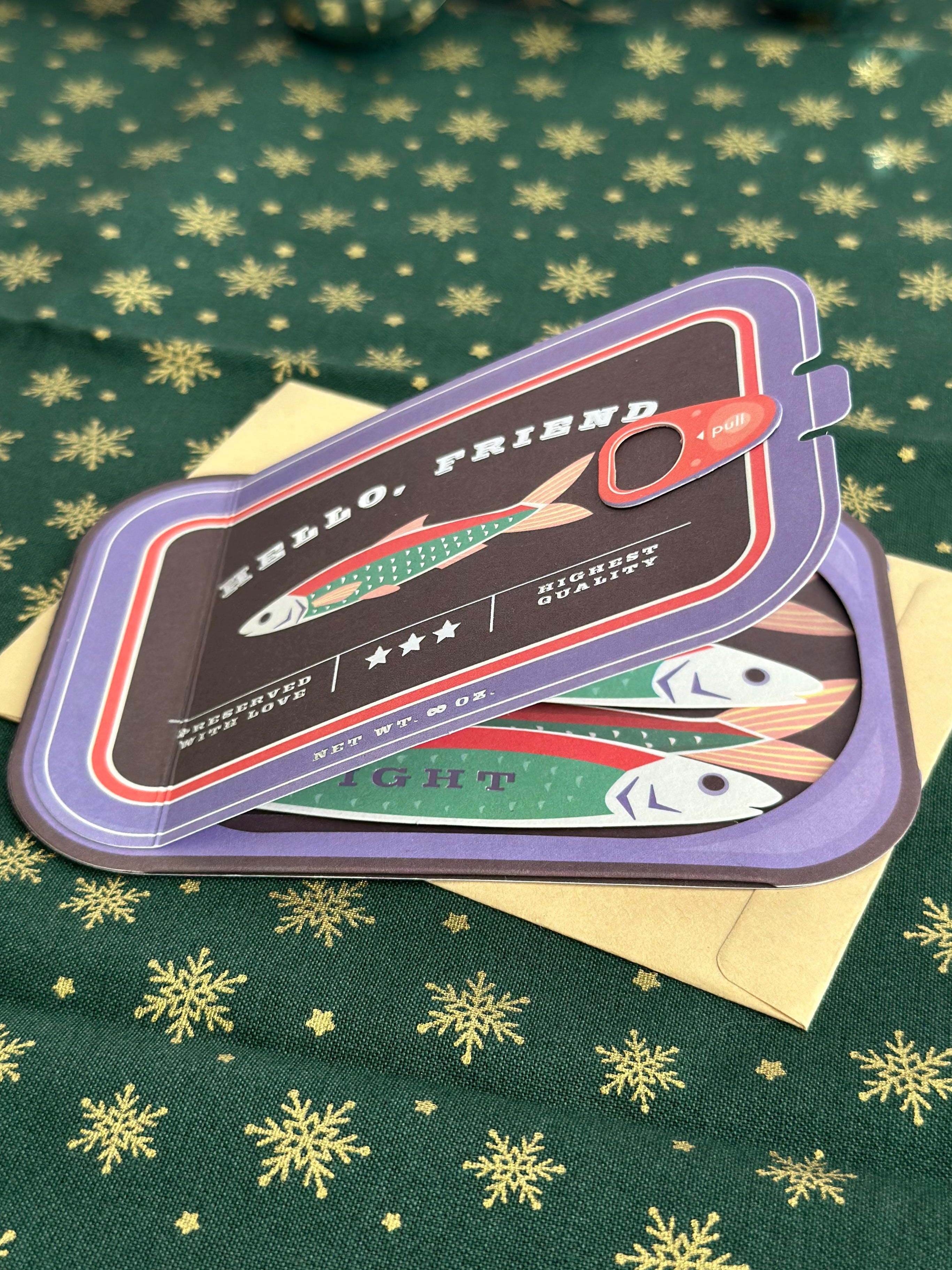 Partially opened sardine-can greeting card with visible pull tab and layered fish artwork, styled on festive green fabric with gold snowflakes.