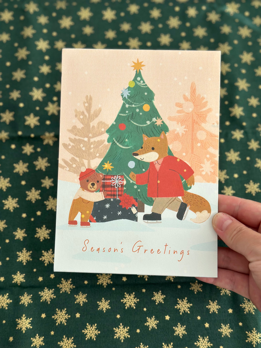 Close view of Season’s Greetings card showing fox in red coat, little bear, and decorated Christmas tree.