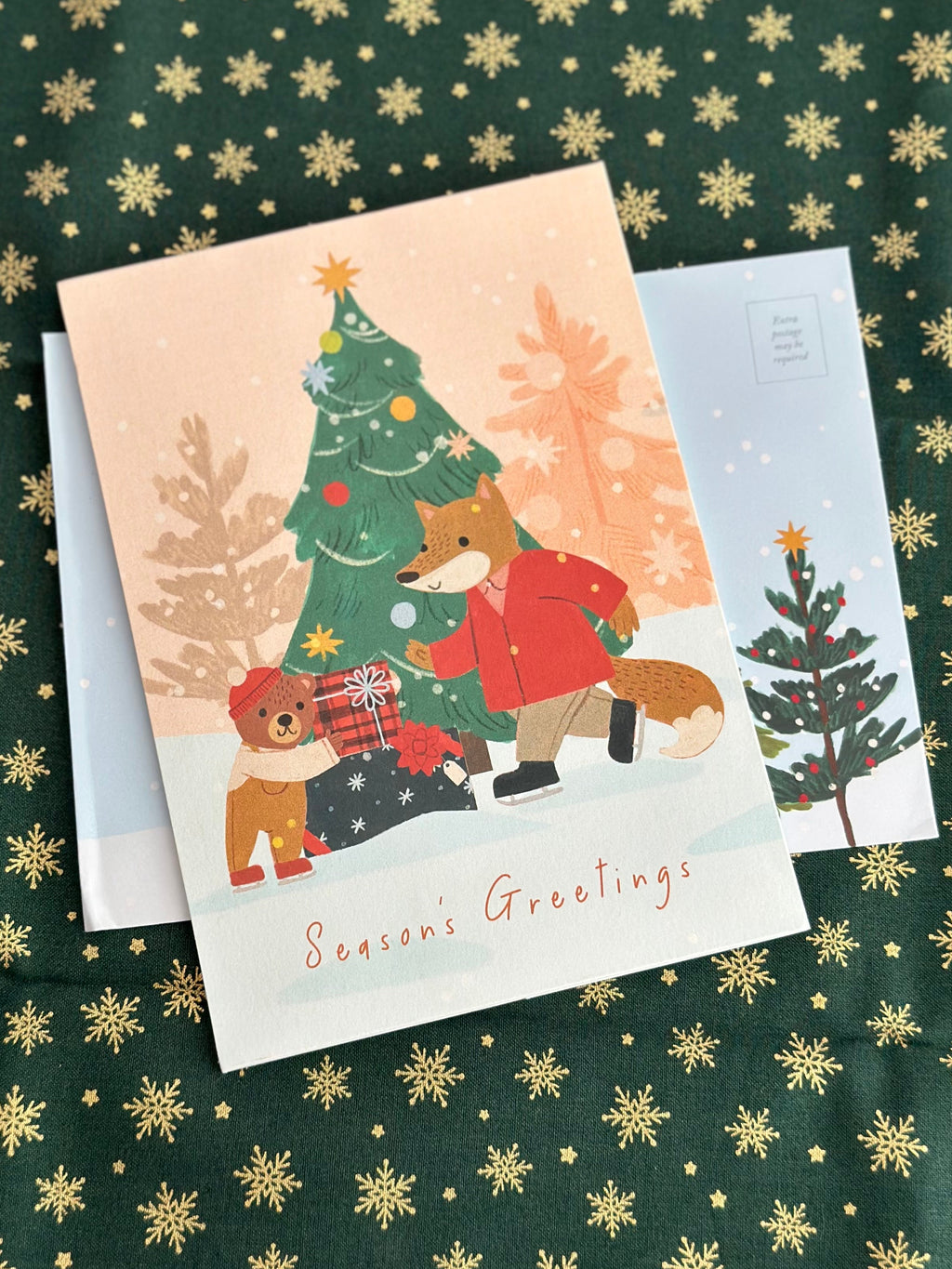 Season’s Greetings card with fox and bear by a Christmas tree, matching illustrated envelope behind, on green fabric with gold snowflakes.