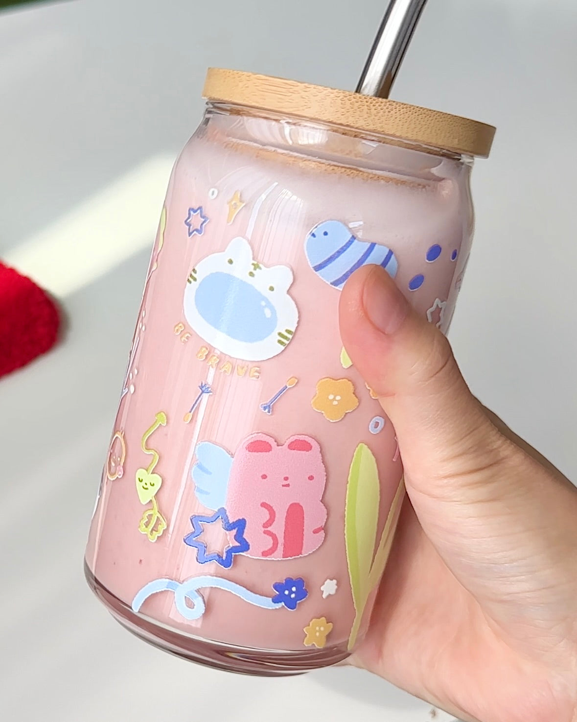 Hand holding a 16 oz Secret Garden glass can with soft affirmation text and pastel illustrations, filled with a pink iced drink and topped with a bamboo lid and glass straw.