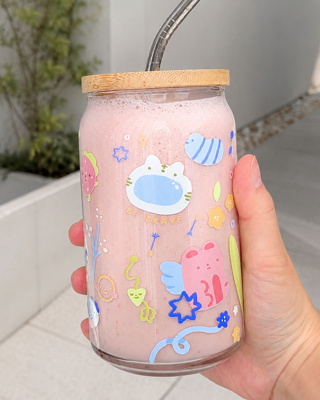 A hand holding a clear glass can-shaped tumbler filled with a pink drink, featuring colorful cartoon animal decals and a bamboo lid with a metal straw, set against a blurred outdoor background.