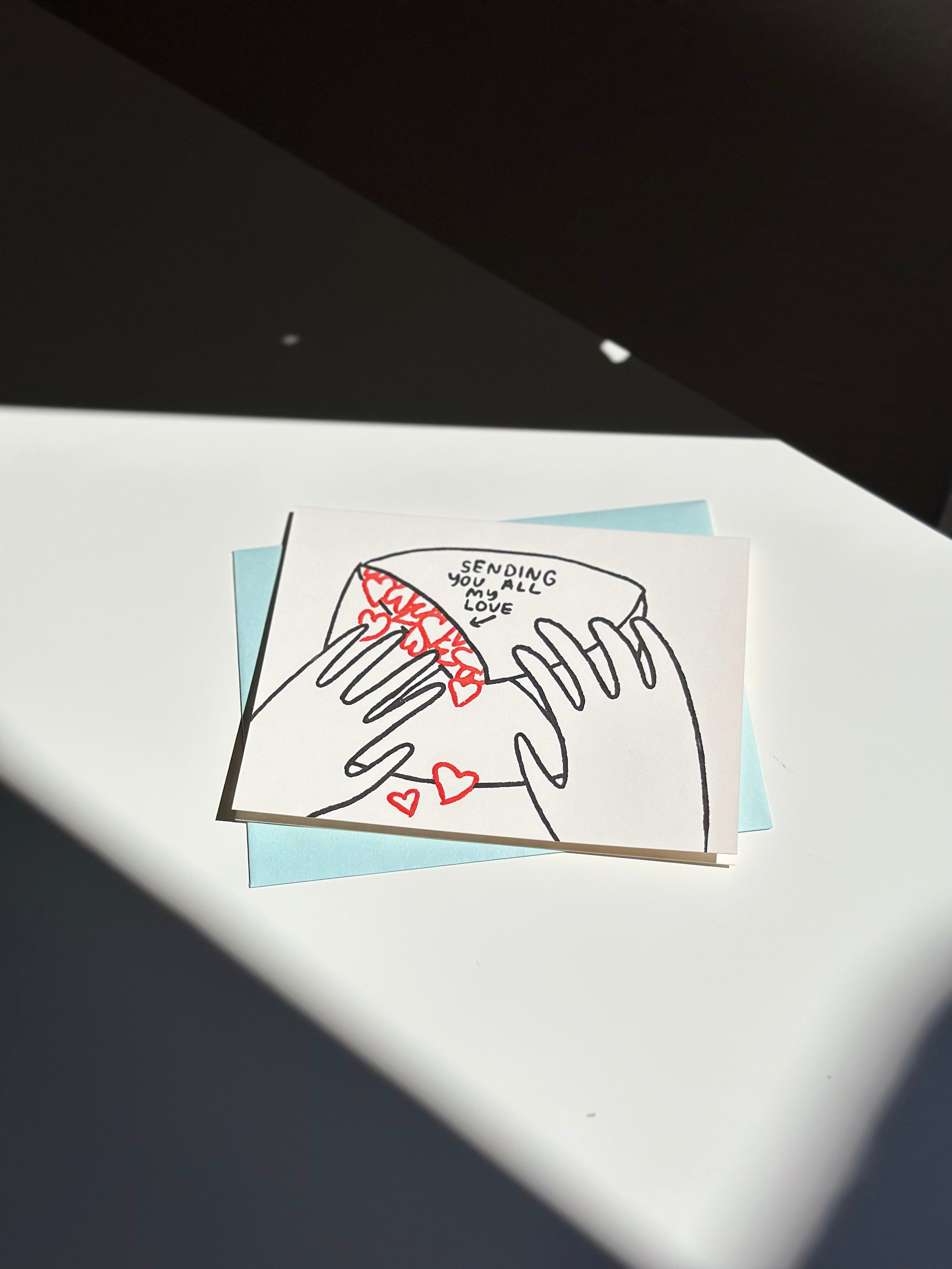 Minimalist love greeting card with illustration of hands holding an envelope spilling out red hearts, captioned ‘Sending you all my love,’ paired with light blue envelope.