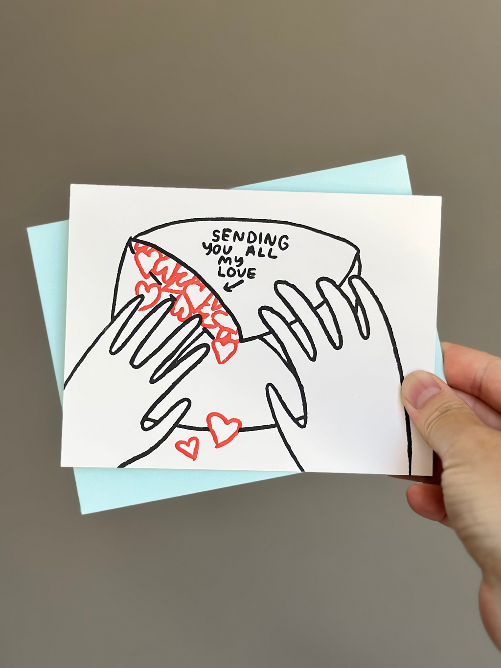 Minimalist love greeting card with illustration of hands holding an envelope spilling out red hearts, captioned ‘Sending you all my love,’ paired with light blue envelope.