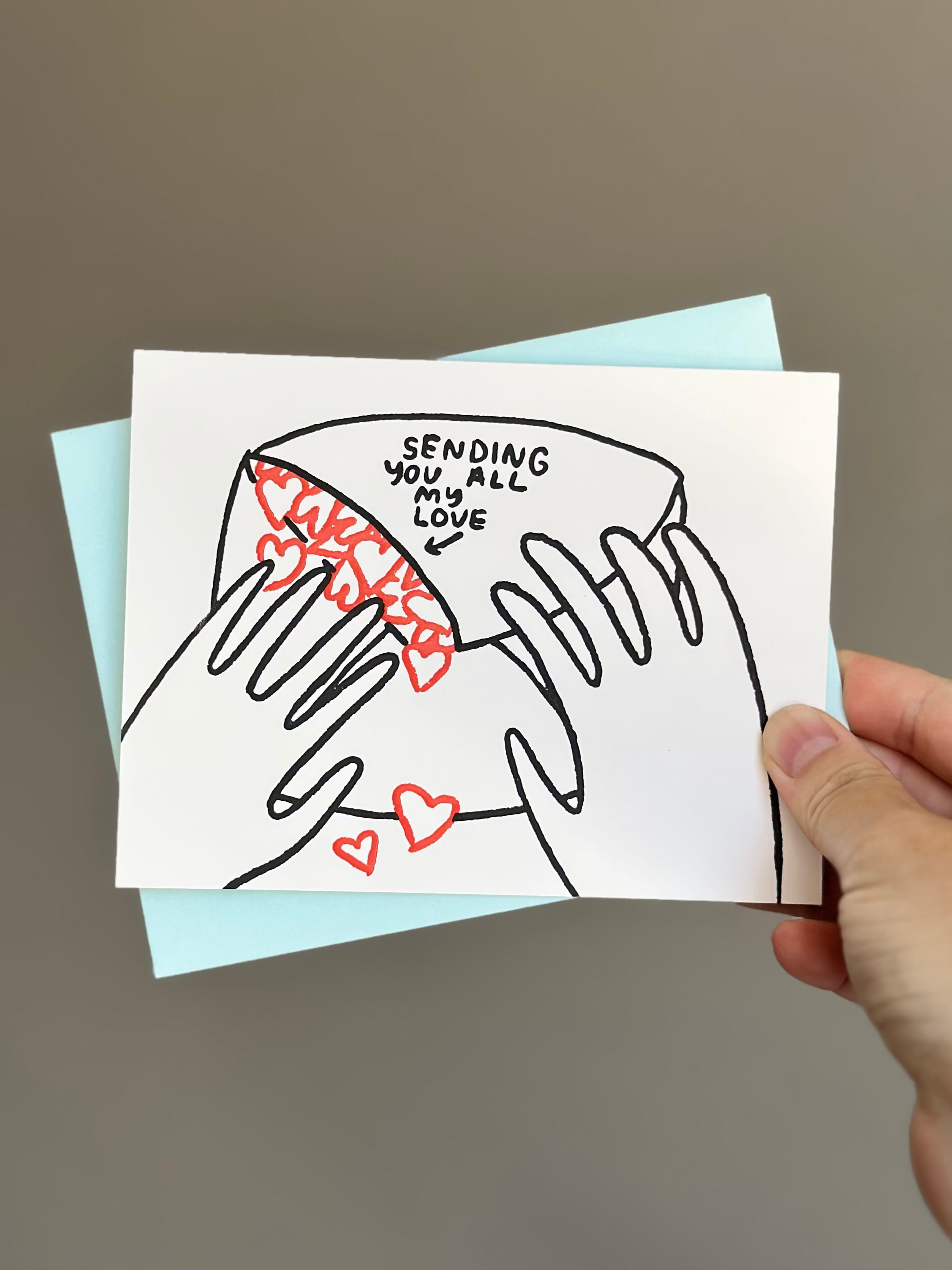 Minimalist love greeting card with illustration of hands holding an envelope spilling out red hearts, captioned ‘Sending you all my love,’ paired with light blue envelope.