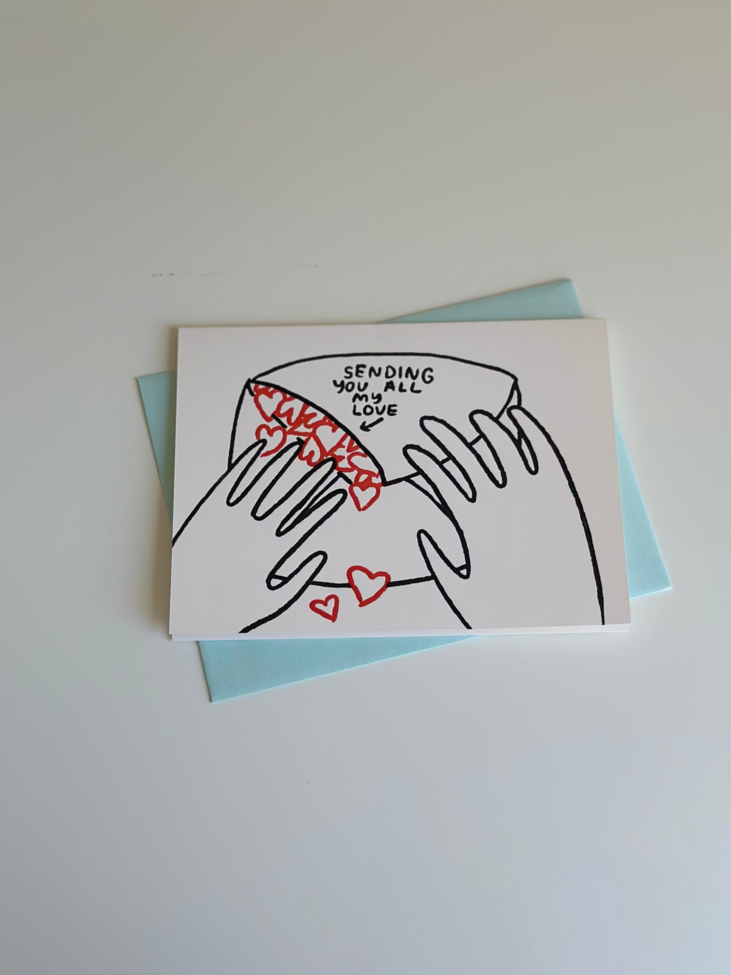 Minimalist love greeting card with illustration of hands holding an envelope spilling out red hearts, captioned ‘Sending you all my love,’ paired with light blue envelope.