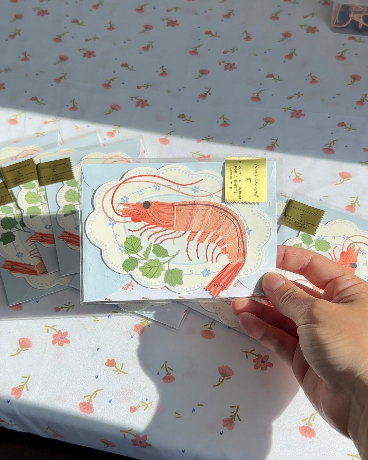 Funny shrimp greeting card in packaging, held over a floral tablecloth.