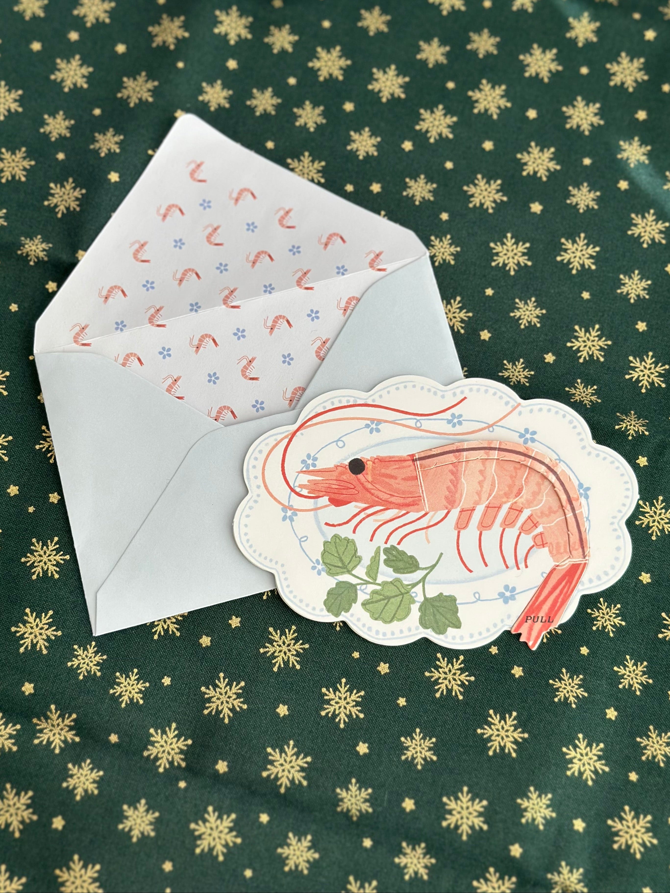 Holiday shrimp greeting card with scalloped edge and blue envelope; shrimp-pattern liner; green fabric with gold snowflakes.