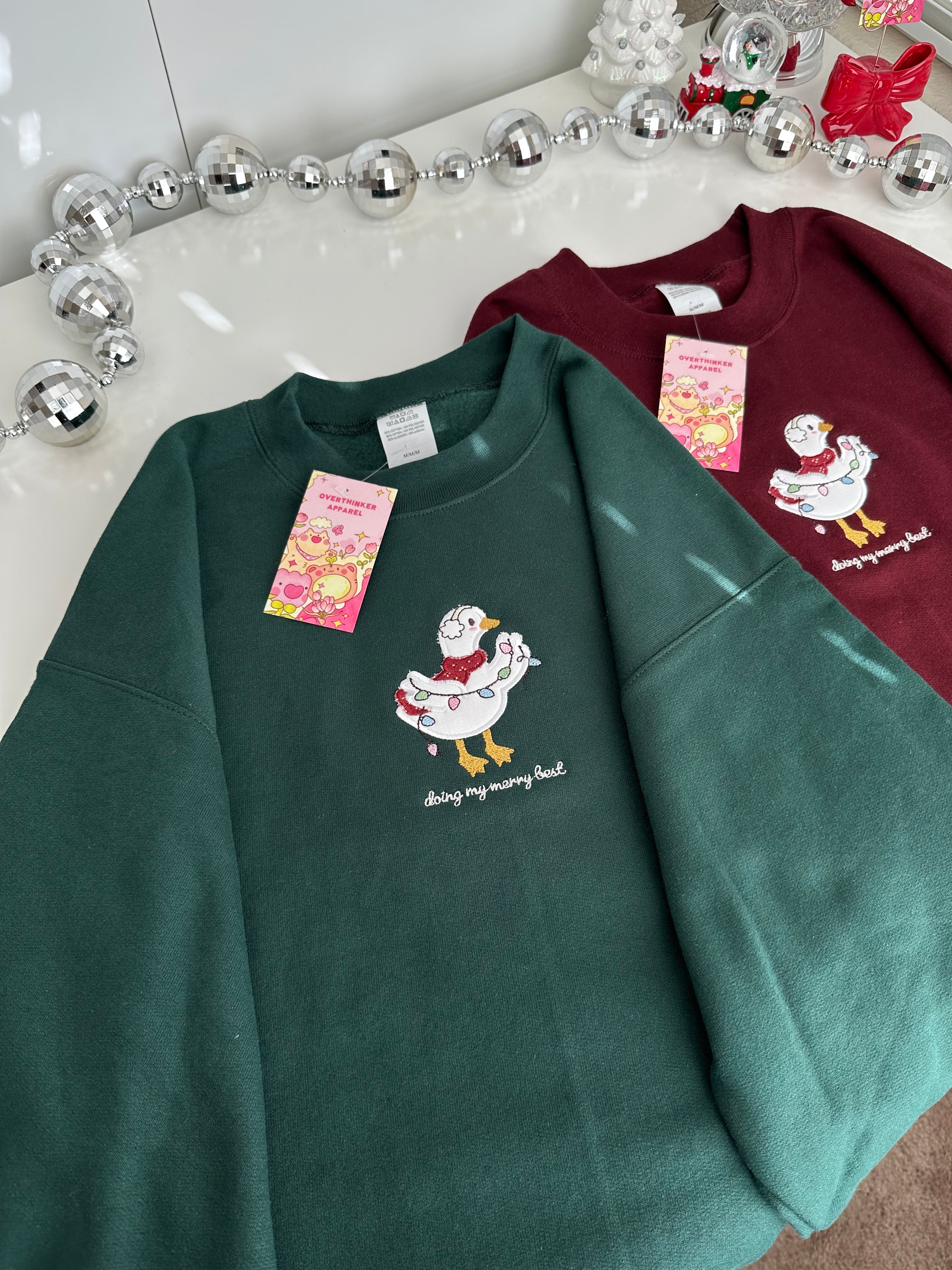 Flat lay of forest green and burgundy oversized holiday sweatshirts featuring an embroidered silly goose design