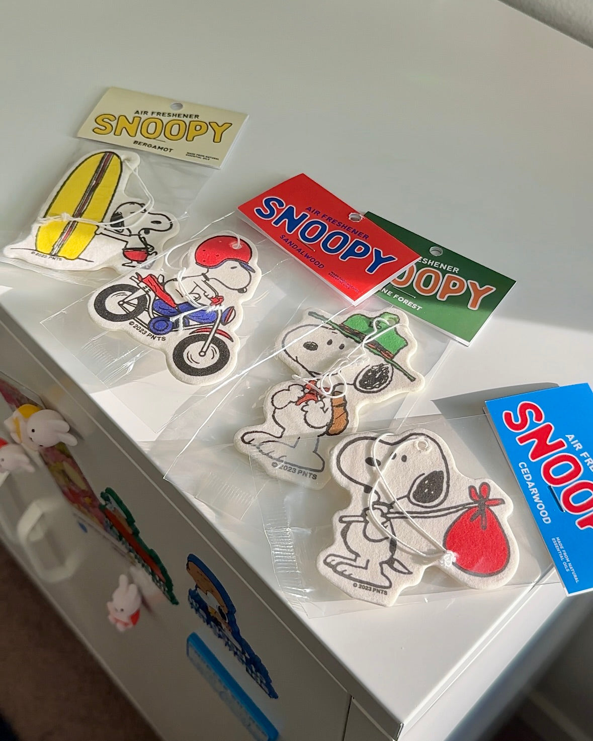 Flatlay of Peanuts air fresheners including Scout Snoopy in green Pine Forest packaging. Perfect for road trips, dorm gifts, or summer vibes.