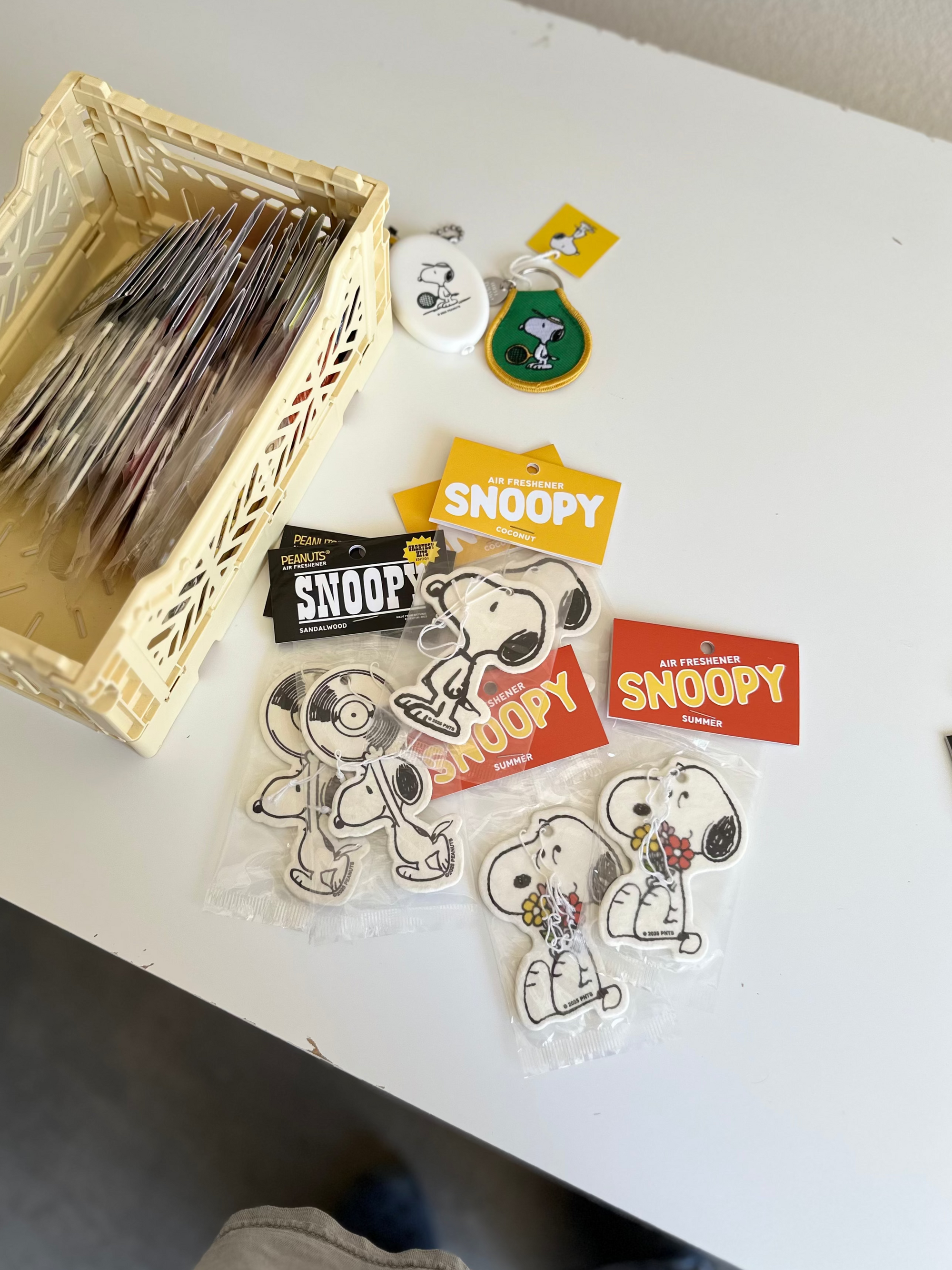 Assorted Snoopy air fresheners in different scents and designs, including coconut, summer, and sandalwood, displayed on a white desk with a beige storage basket and Peanuts keychains.