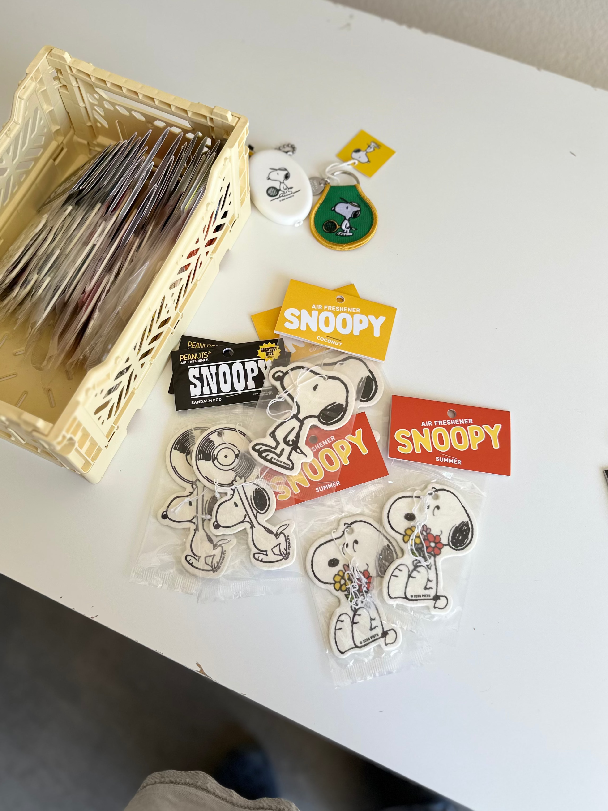 Assorted Snoopy air fresheners in different scents and designs, including coconut, summer, and sandalwood, displayed on a white desk with a beige storage basket and Peanuts keychains.