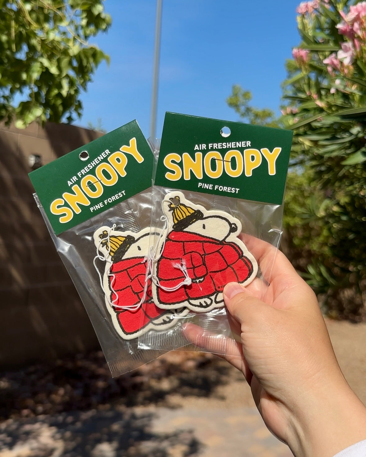 Hand holding two Snoopy puffer jacket air fresheners in Pine Forest scent against a sunny blue sky background.