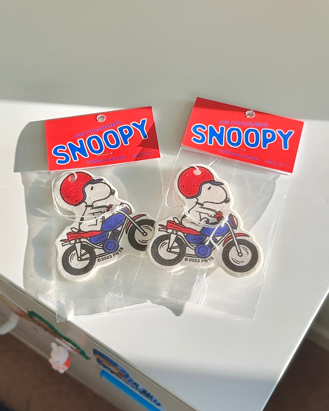 Flatlay of two Snoopy motorcycle air fresheners in sandalwood scent on a white surface. Cute retro car accessories for Peanuts fans and gift shoppers.