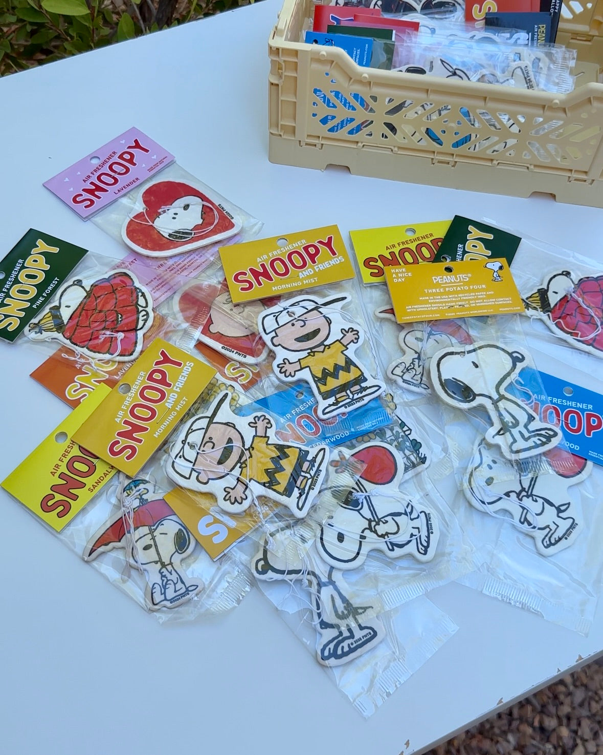 Flat lay of assorted Snoopy and Charlie Brown air fresheners in various scents, arranged on a white table next to a beige crate.