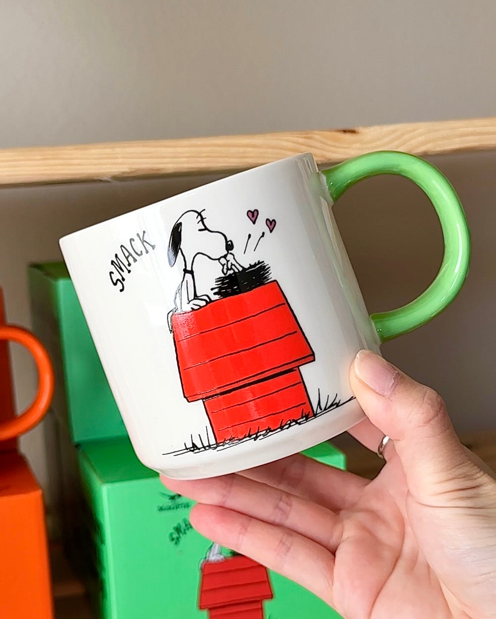 White ceramic Snoopy coffee mug featuring Snoopy on his red doghouse with floating hearts and the phrase “Be Kind to All Kinds,” Peanuts mug with green handle.