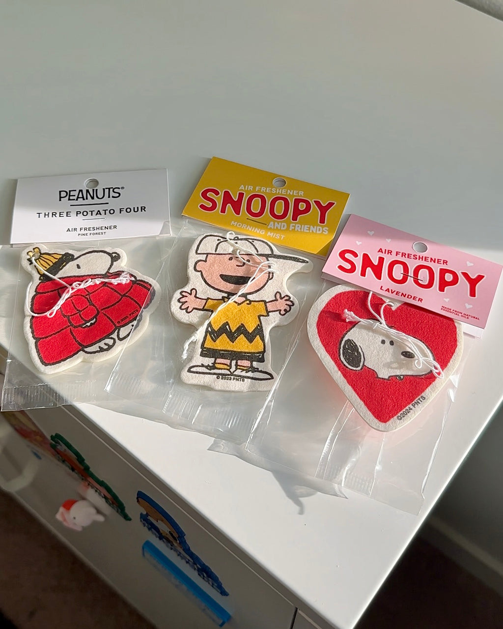 Three Peanuts® air fresheners featuring Snoopy in a red puffer jacket, Charlie Brown, and Snoopy in a heart design, in scents Pine Forest, Morning Mist, and Lavender.