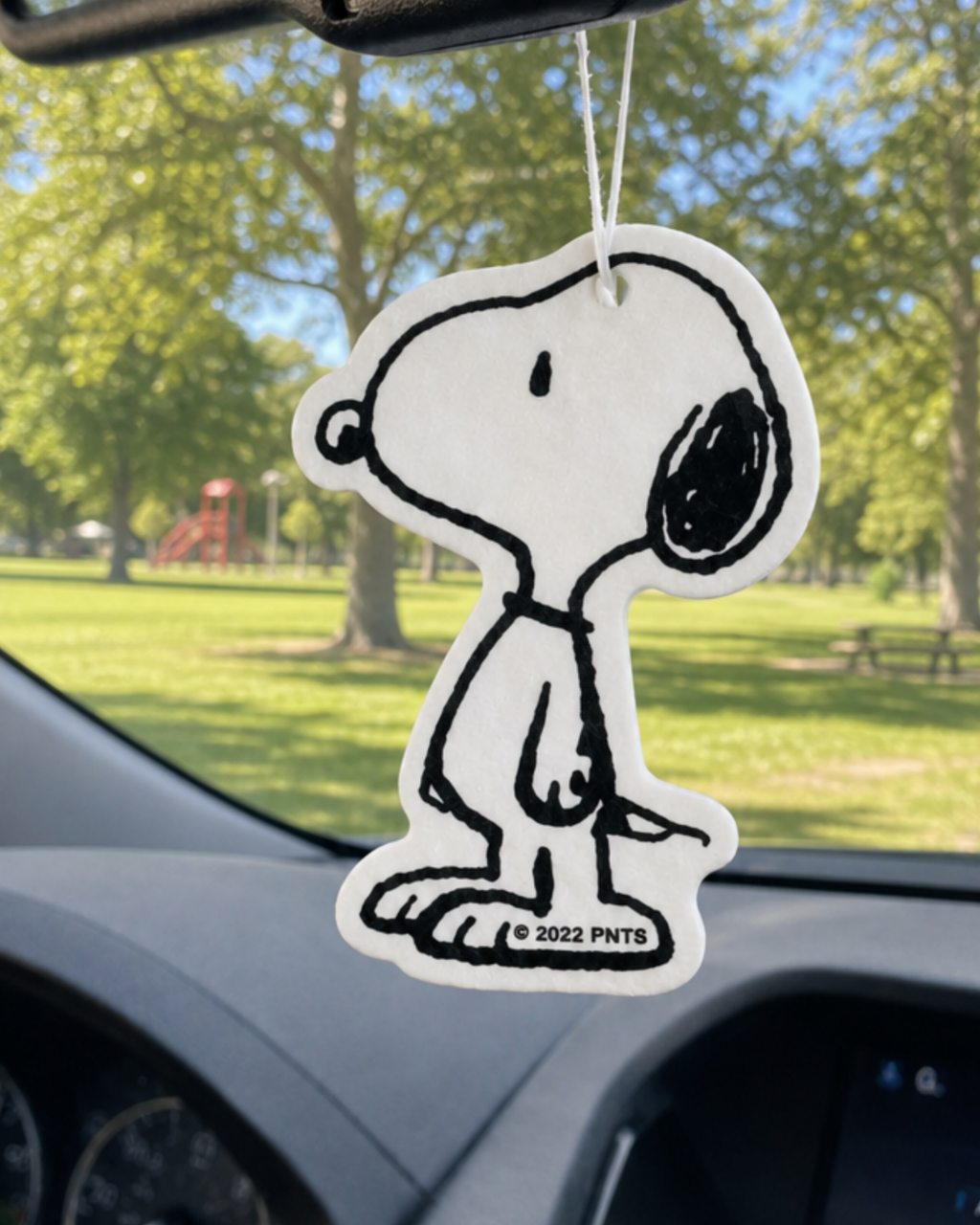 Classic black and white Snoopy air freshener hanging from a car mirror with a green park and playground in the background.