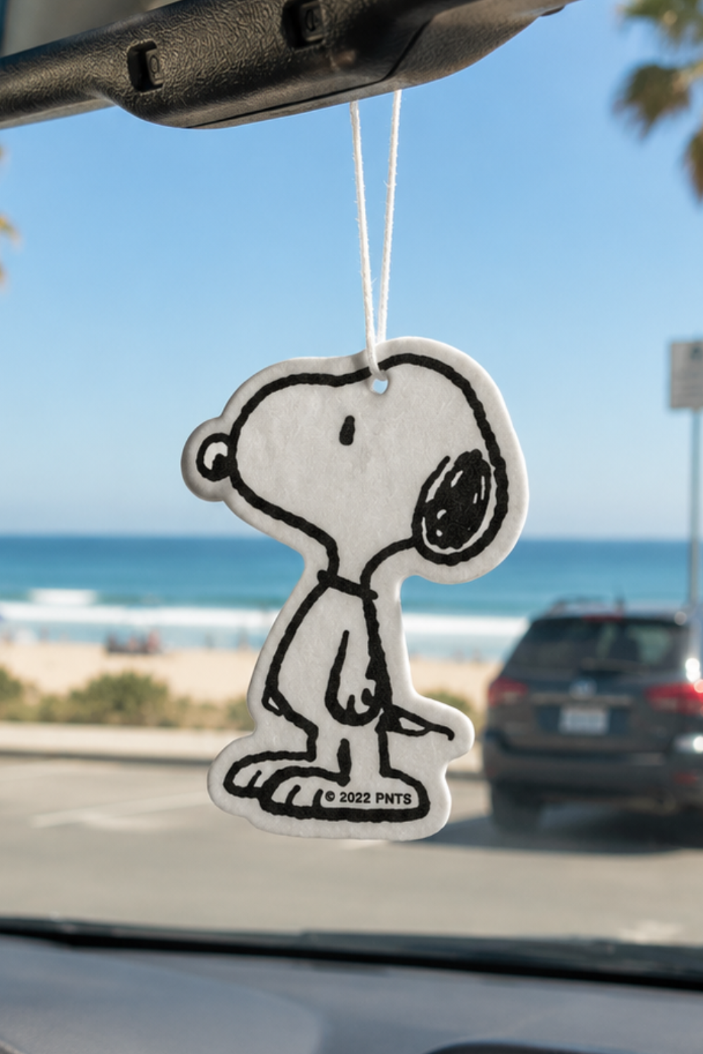 Classic black-and-white Snoopy car air freshener hanging from a rearview mirror with a beach and ocean view in the background.