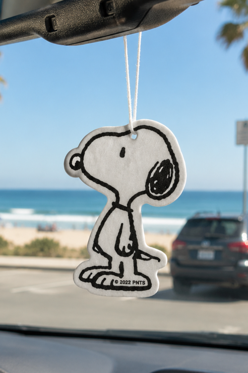 Classic black-and-white Snoopy car air freshener hanging from a rearview mirror with a beach and ocean view in the background.