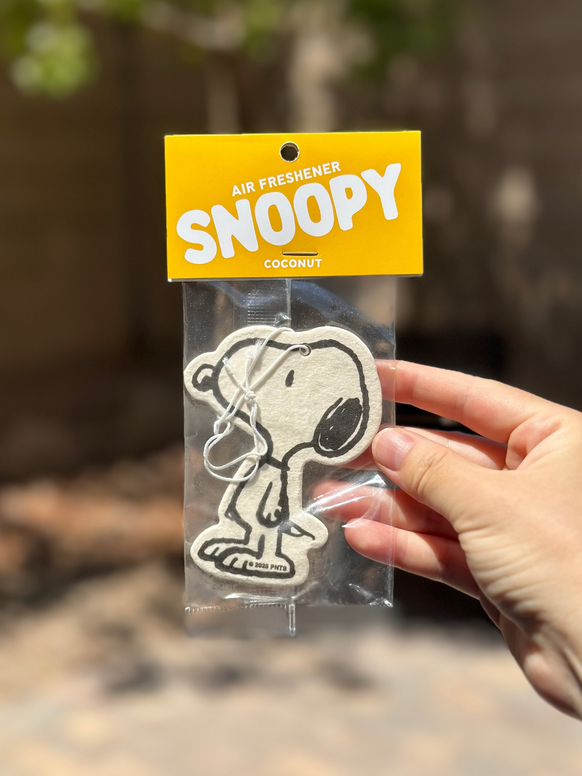Close-up of a single Snoopy coconut air freshener in packaging, held outdoors against a blurred leafy background.