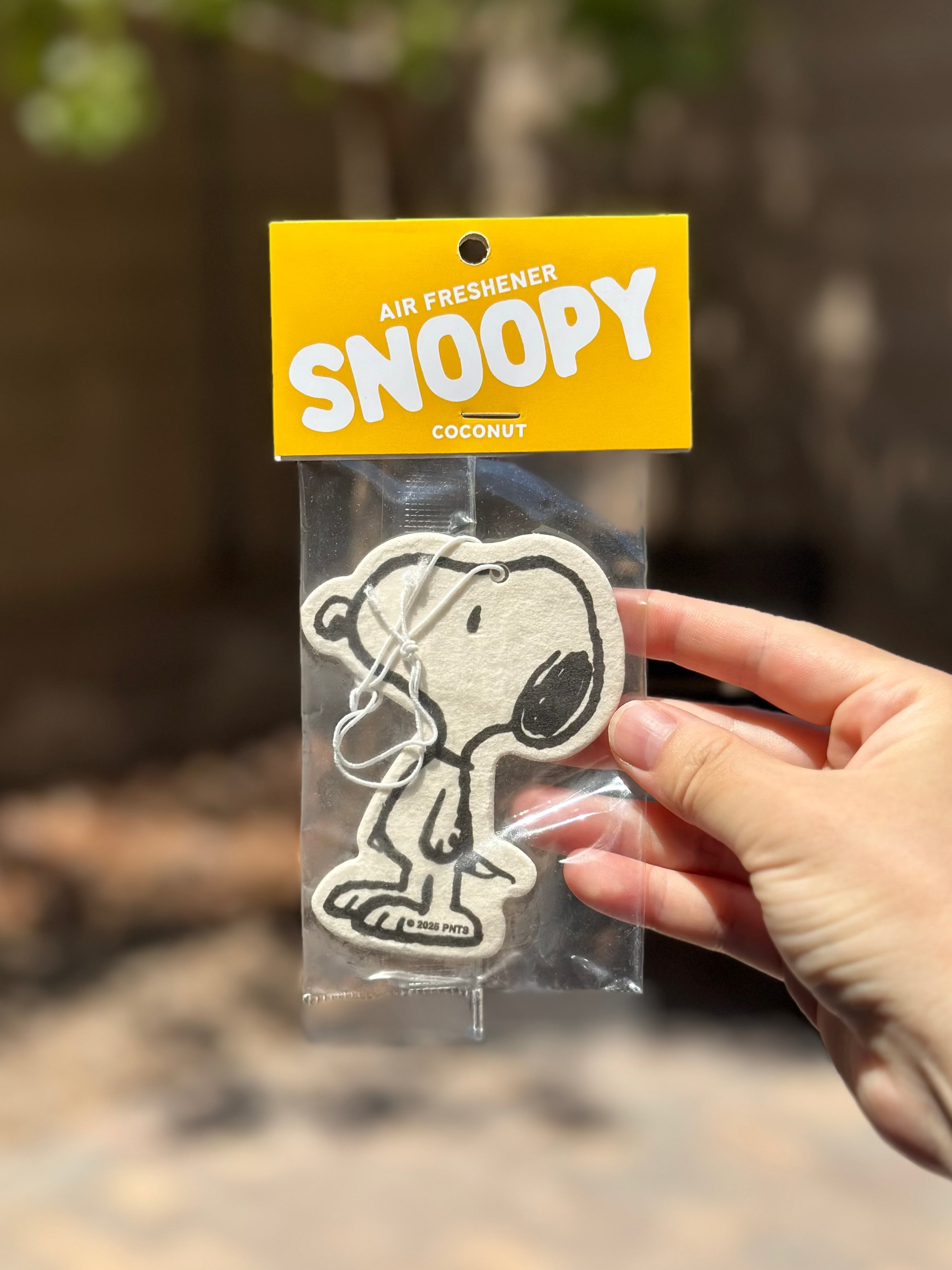 Close-up of a single Snoopy coconut air freshener in packaging, held outdoors against a blurred leafy background.