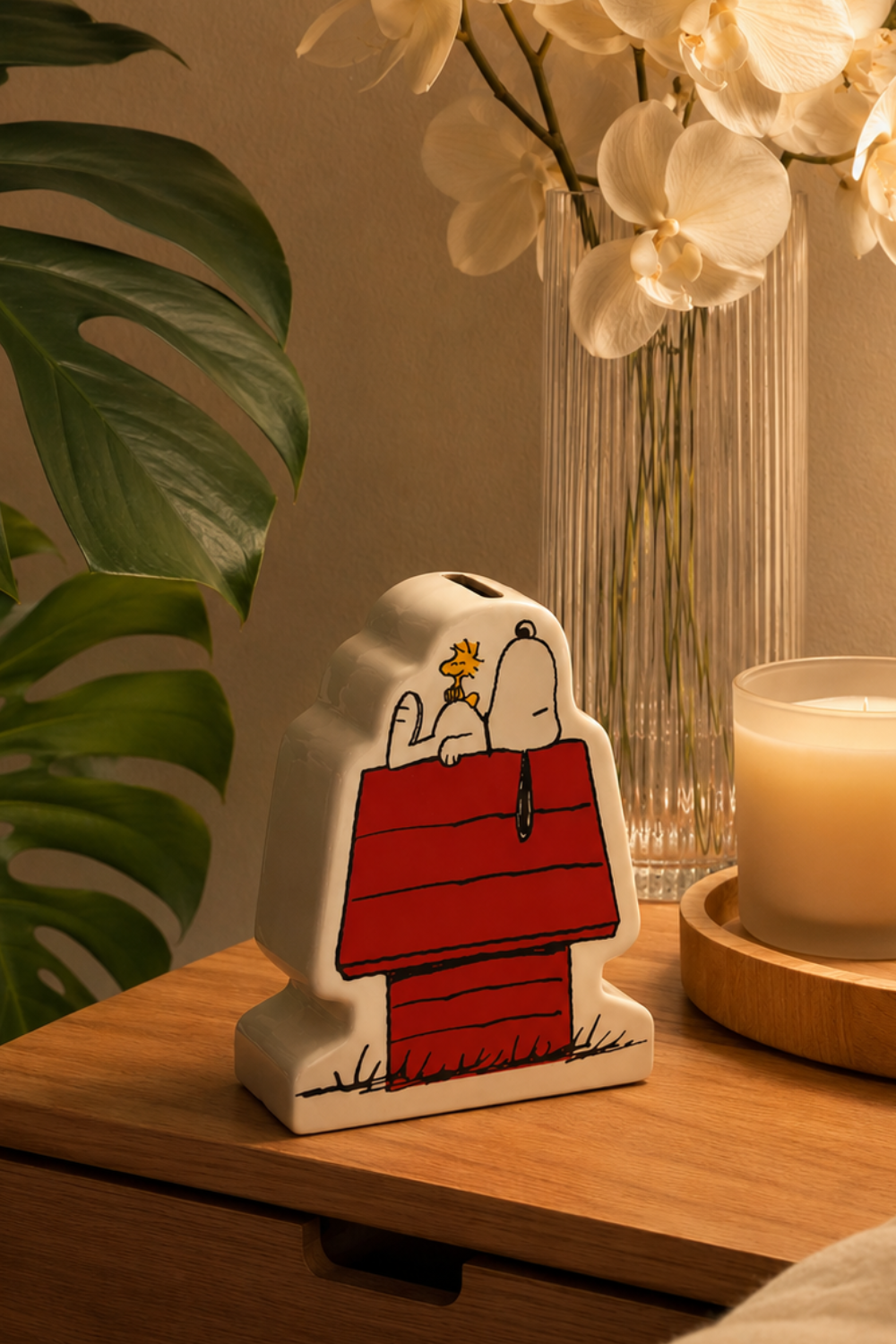 Snoopy and Woodstock ceramic coin bank glowing beside a lit candle on a wooden nightstand, a cozy Peanuts bedroom decor accent.
