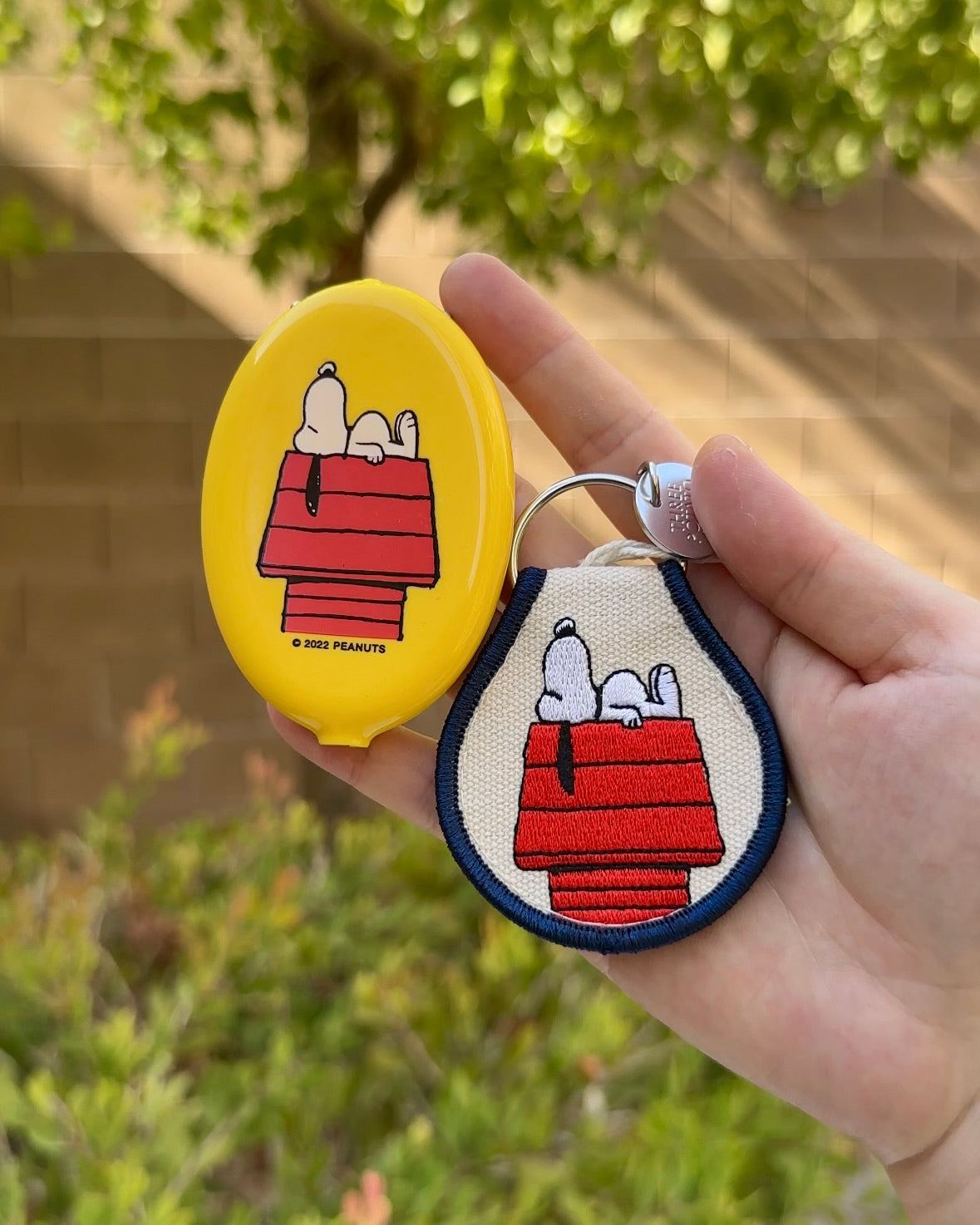 Hand holding Snoopy yellow coin pouch and embroidered Snoopy keychain, both featuring Snoopy sleeping on red doghouse, Peanuts® licensed accessories.