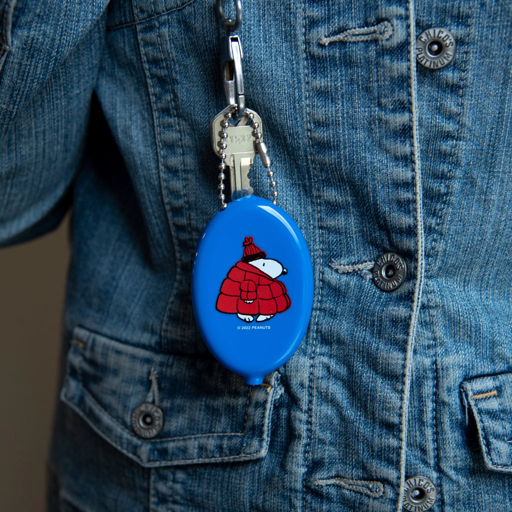 Snoopy coin pouch keychain clipped to a denim jacket. Features Snoopy in a red puffer coat and beanie, attached with a silver ball chain and key — a cozy winter-ready Peanuts accessory.
