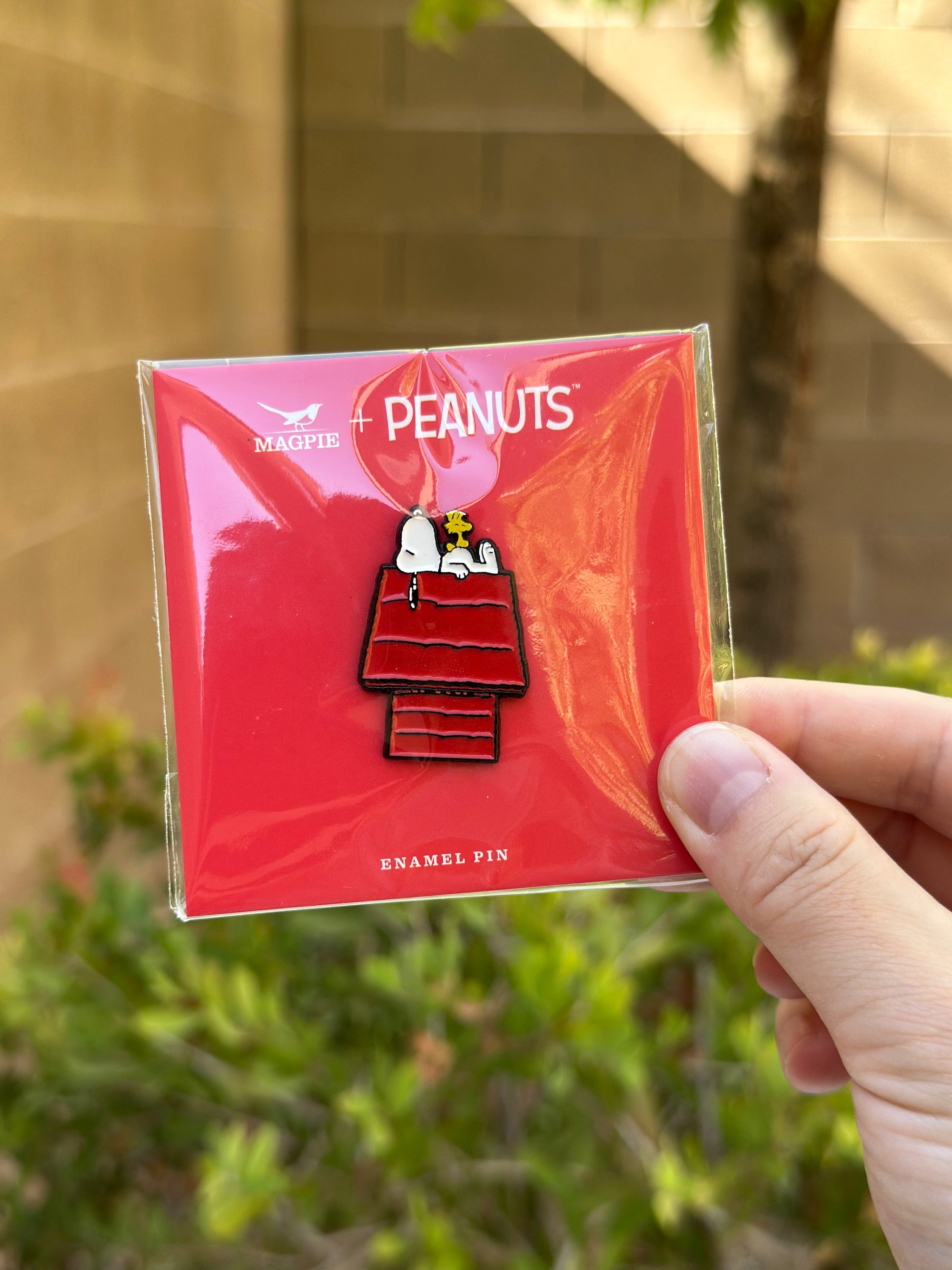 Snoopy and Woodstock enamel pin on red Peanuts® card, held outdoors with greenery in background