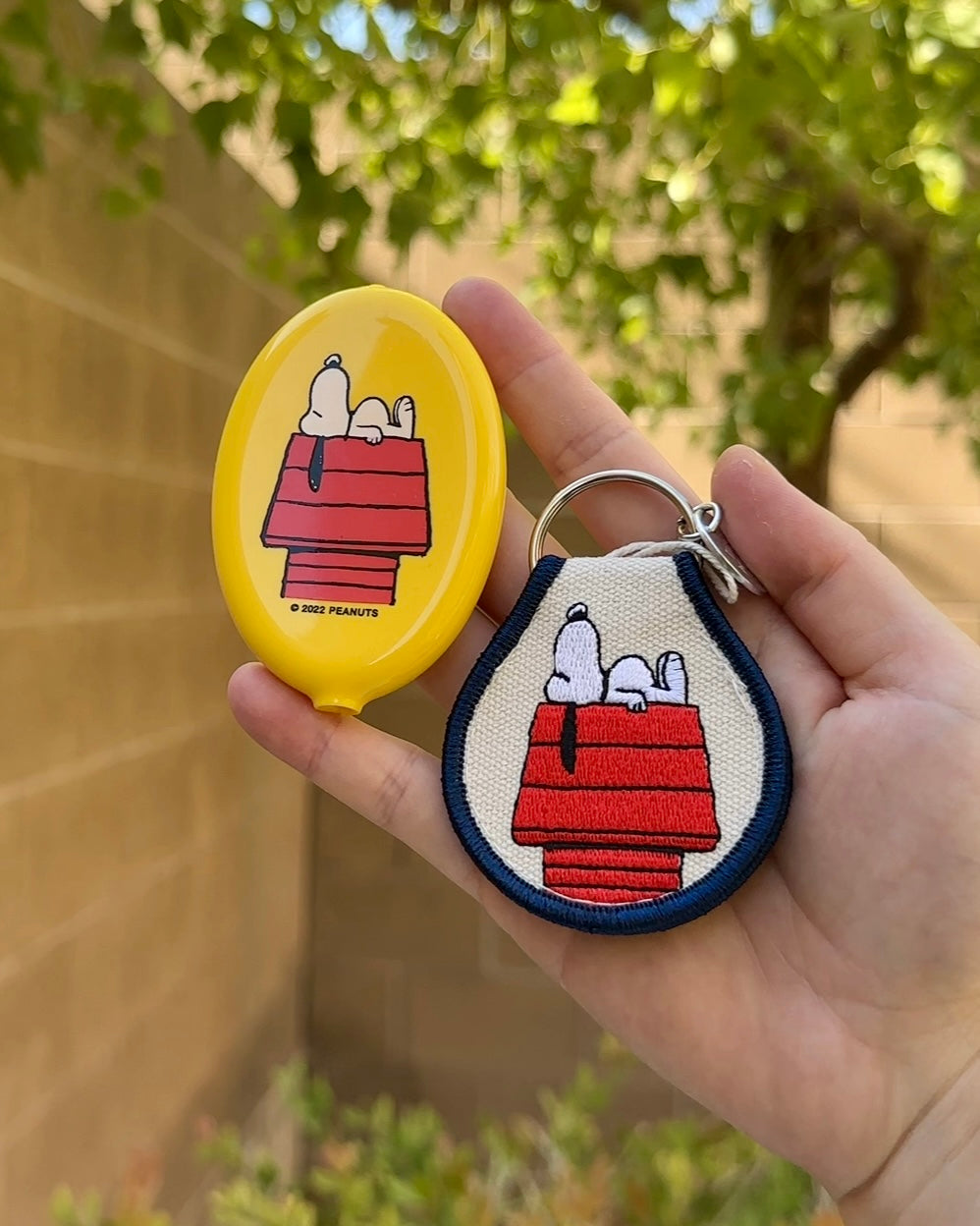Hand holding Snoopy Peanuts embroidered doghouse keychain and yellow coin pouch featuring Snoopy sleeping on his red doghouse.