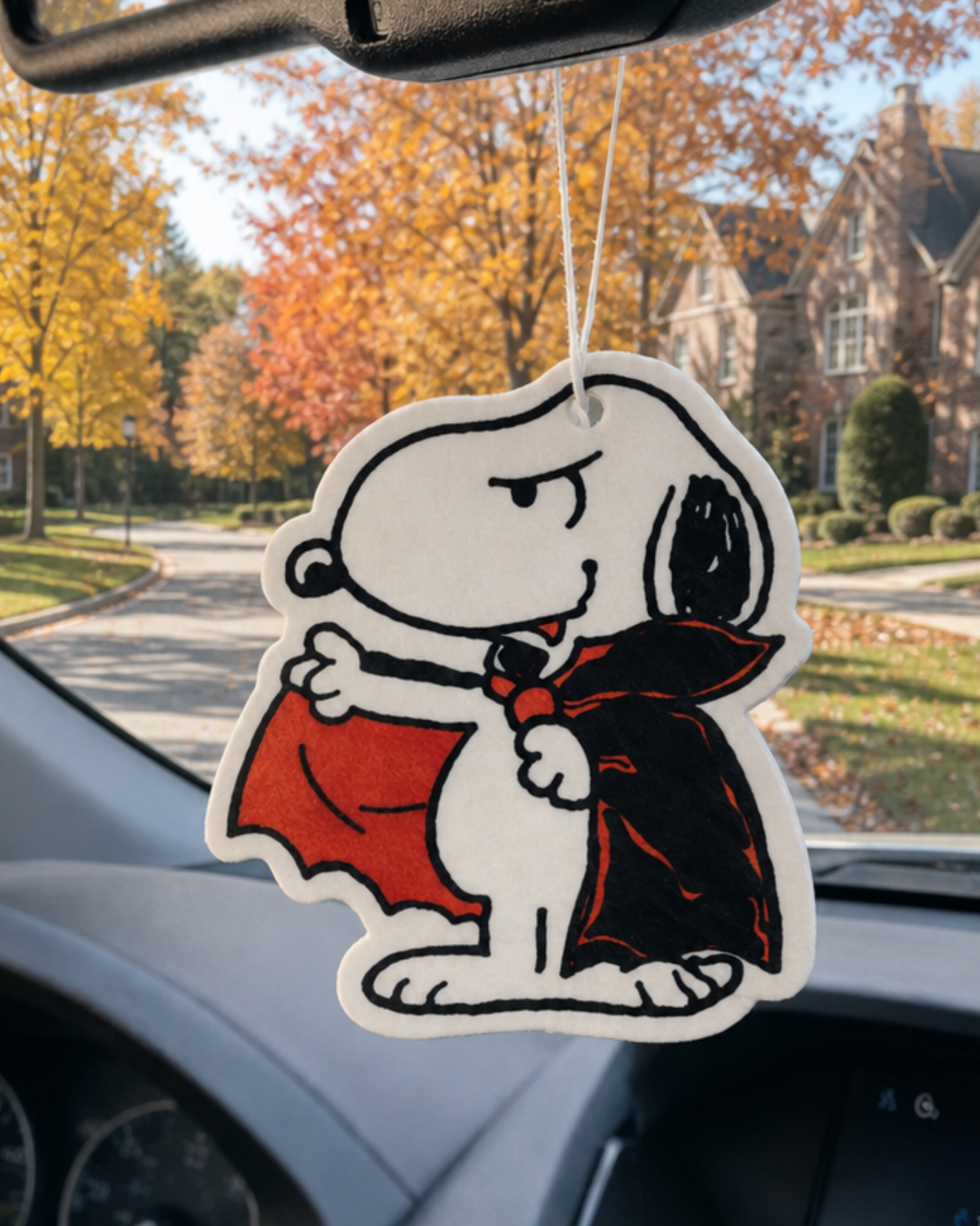 Snoopy Dracula air freshener in a black and red cape, hanging from a car mirror on a suburban street lined with fall trees.