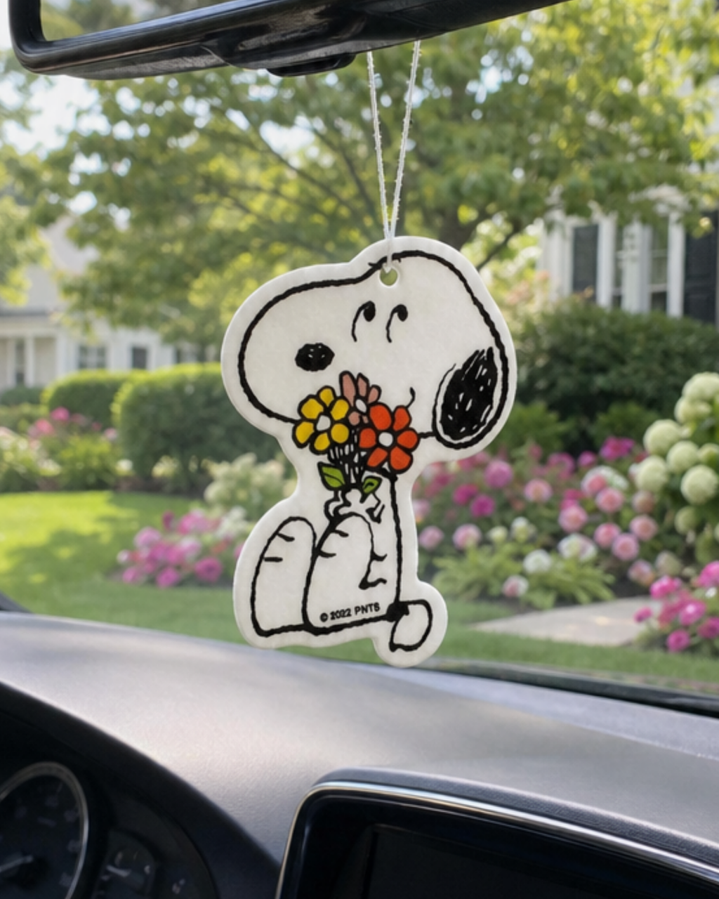 Full color Snoopy air freshener holding a bouquet of flowers, hanging from a car mirror with pink and white garden blooms in the background.
