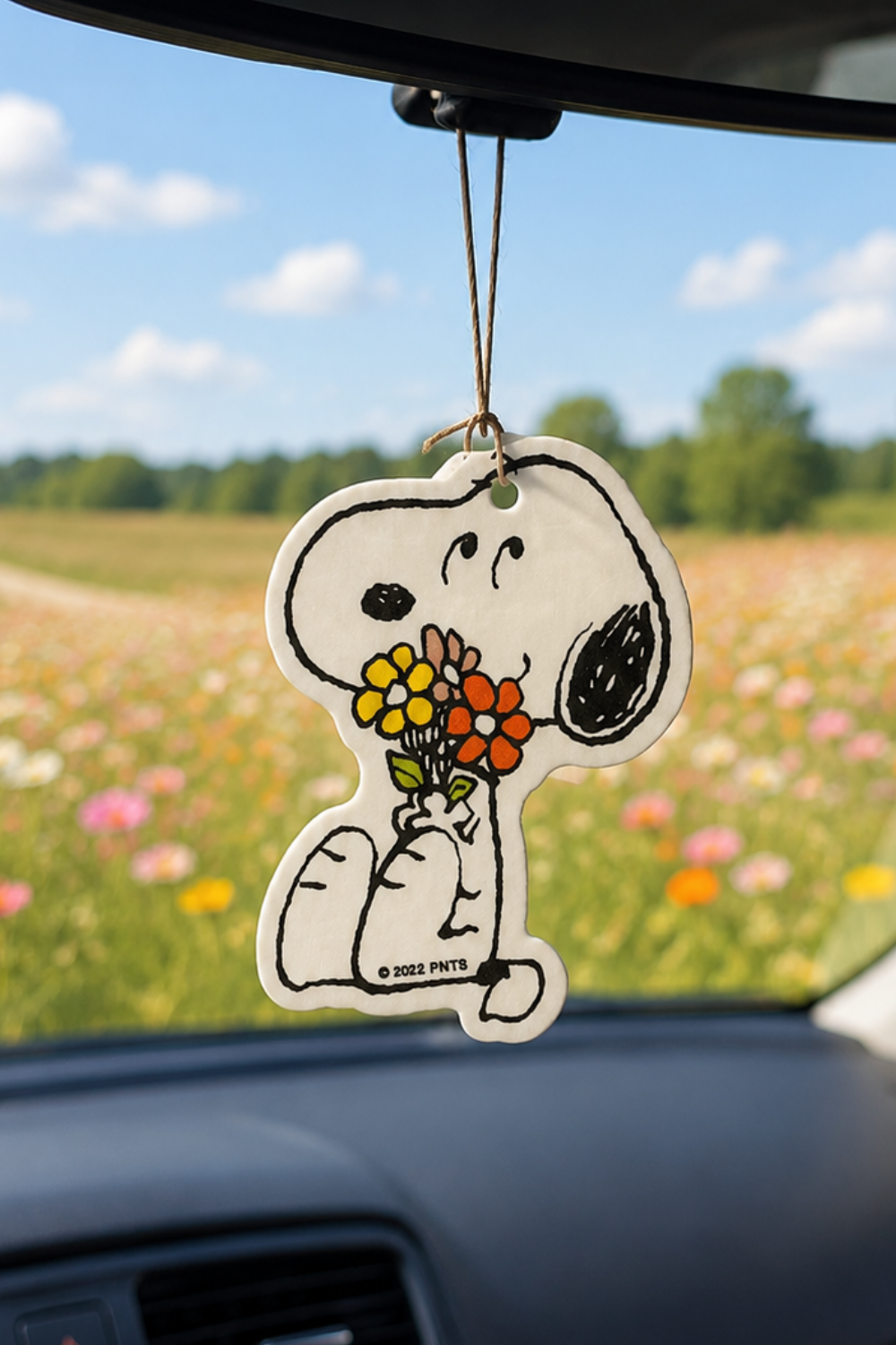 Snoopy car air freshener holding a bouquet of orange and yellow flowers, hanging in a car overlooking a wildflower field.