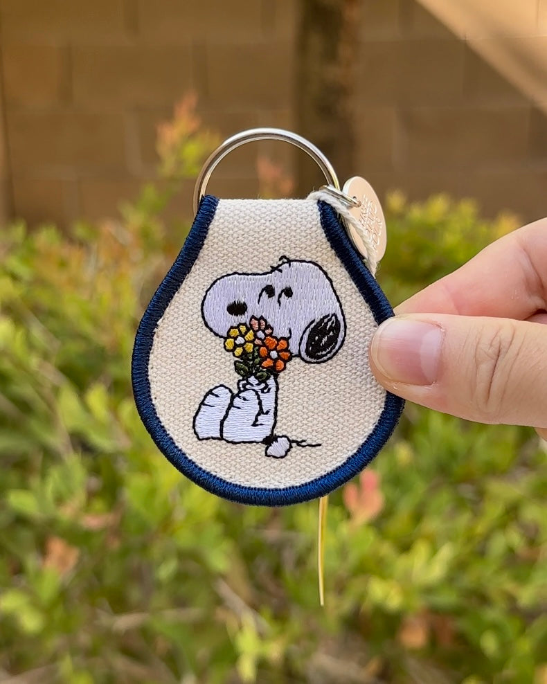 Close-up of embroidered Snoopy holding flowers keychain with navy blue border, Peanuts® collectible patch.