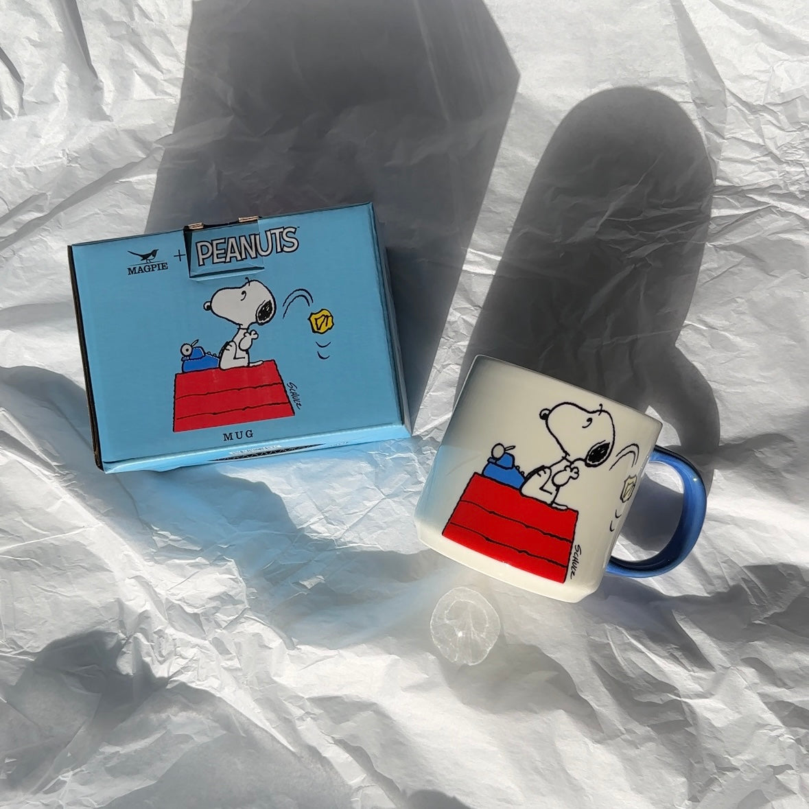 Snoopy mug and blue gift box with Peanuts® branding on crinkled paper background