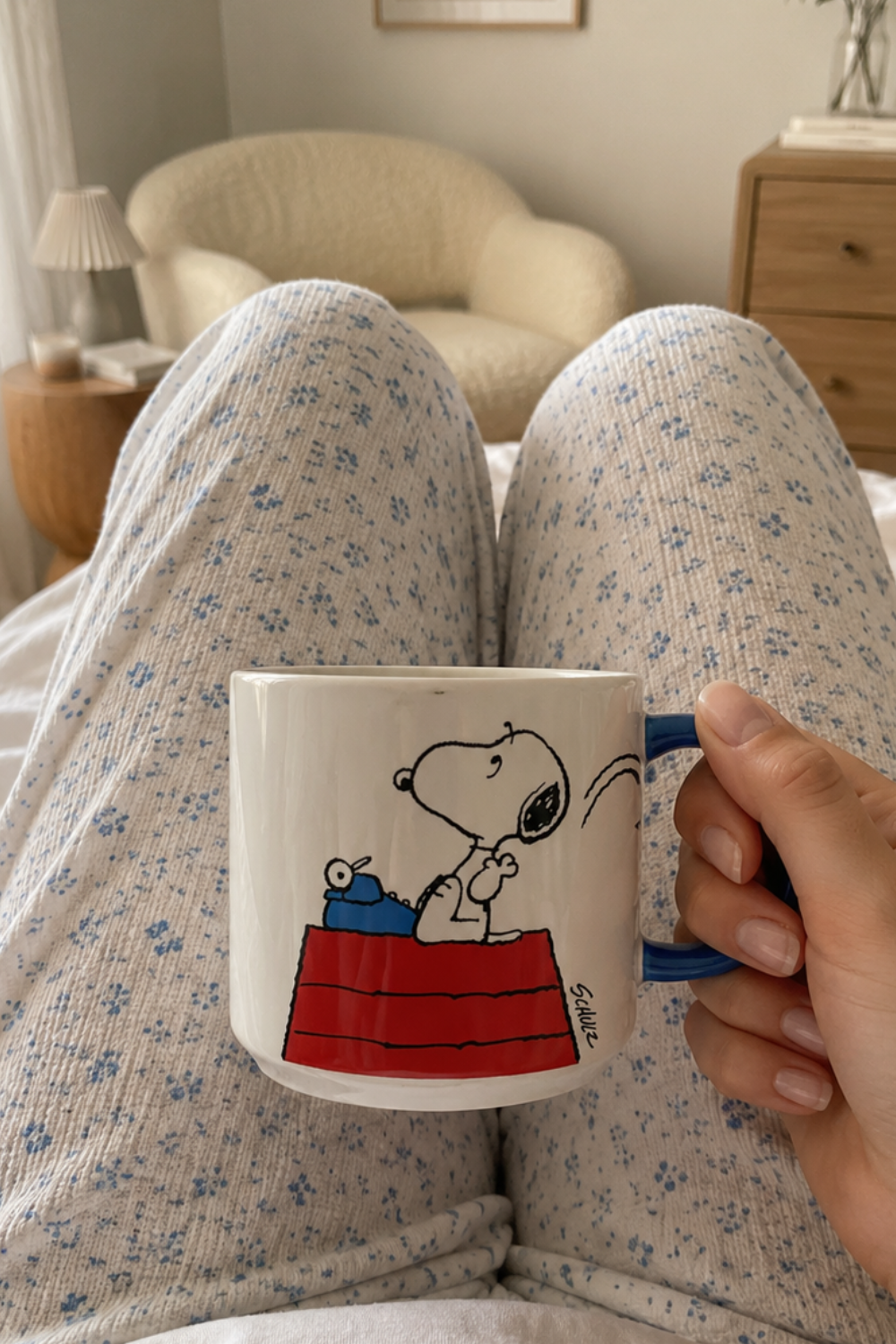 Woman relaxing in bed holding a Snoopy Genius at Work ceramic coffee mug with blue handle in a cozy bedroom morning setting.
