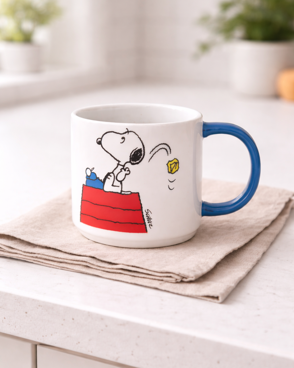 Snoopy Genius at Work Peanuts ceramic mug with blue handle on a neutral linen cloth in a bright kitchen, minimalist coffee mug for cozy mornings and creative desks