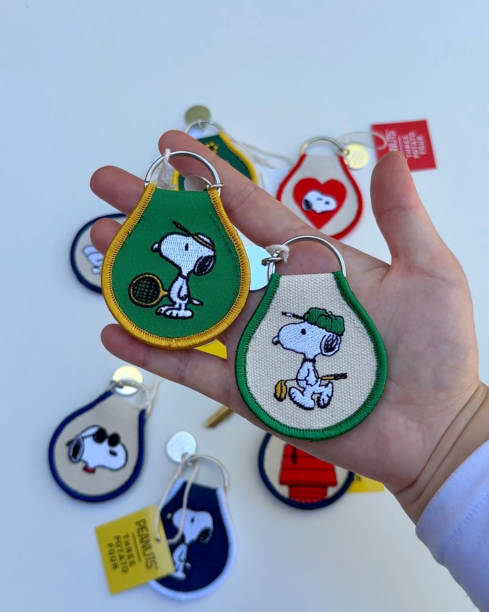 Snoopy Peanuts embroidered golf and tennis keychains held in hand, with colorful Peanuts keychain collection displayed in background.