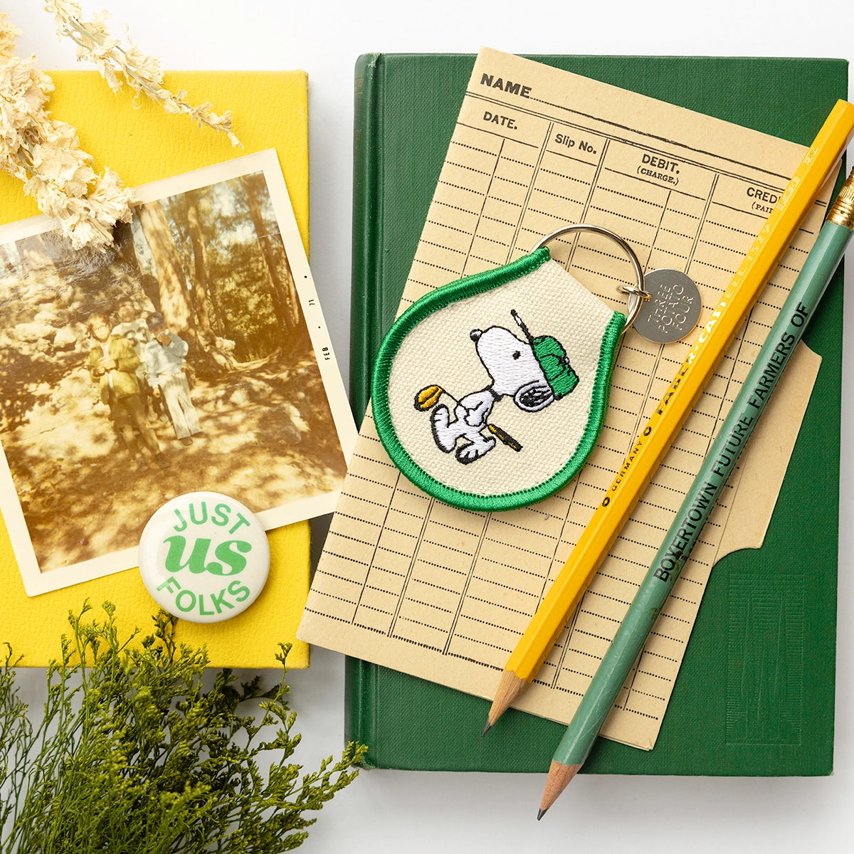 Flat lay featuring a Snoopy golf keychain on vintage library cards and books. Embroidered design with green trim shows Snoopy in a cap holding a golf club. A nostalgic Peanuts accessory with charming retro details.
