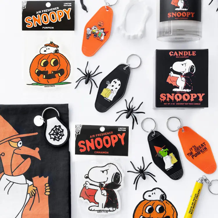 Flat lay of Snoopy Halloween merchandise including air fresheners, candles, keychains, and spooky decorations. Features multiple Peanuts characters in themed packaging — perfect for Halloween gifting or collectors.

