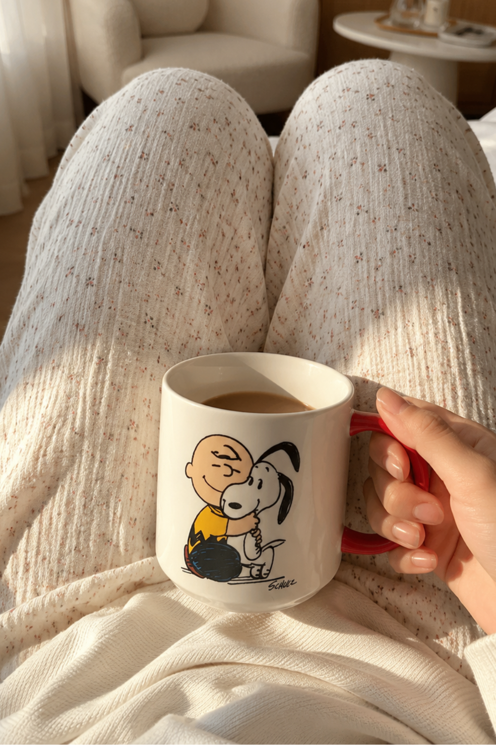 Cozy bedroom morning scene featuring a Snoopy Happiness Is a Warm Puppy coffee mug held above soft cream pajamas with coffee inside.