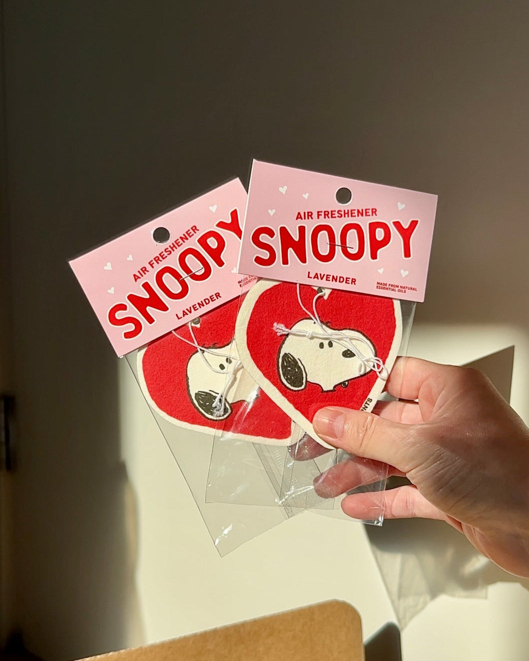 Two heart-shaped Snoopy lavender air fresheners held in hand under warm sunlight. Cute car decor with a soft pink Peanuts-themed label.