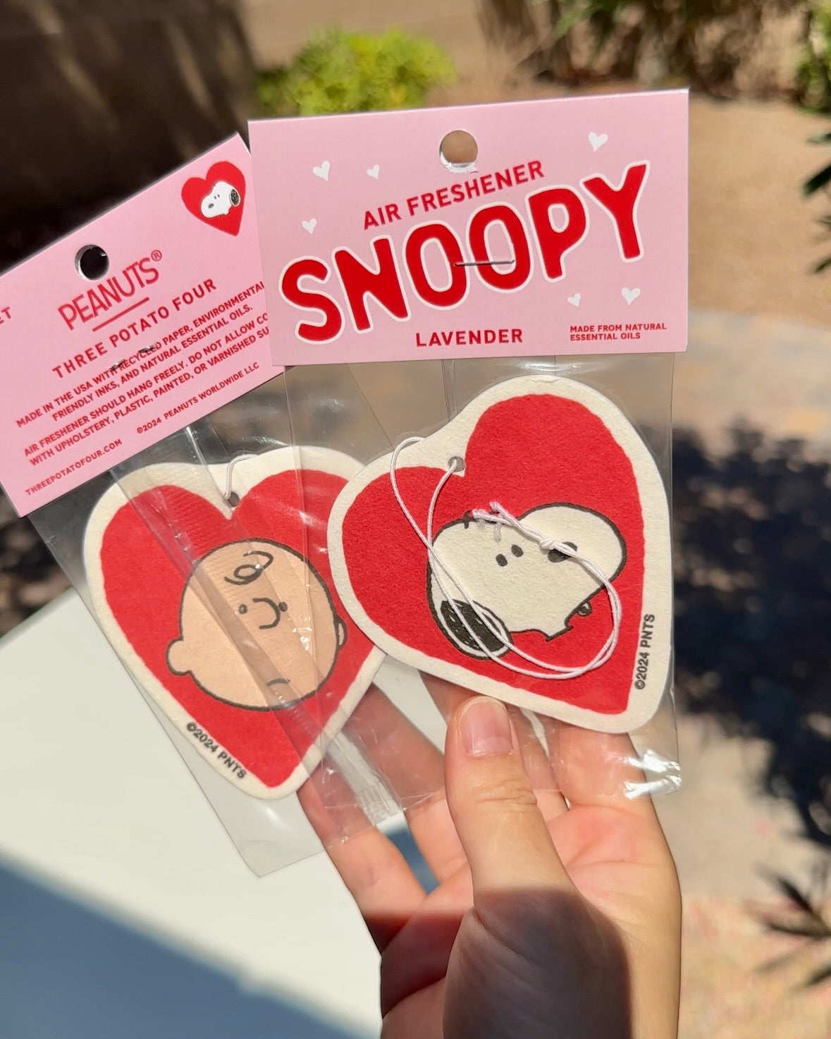 Close-up of hand holding two heart-shaped Peanuts® air fresheners, one with Snoopy and one with Charlie Brown, in lavender scent packaging.