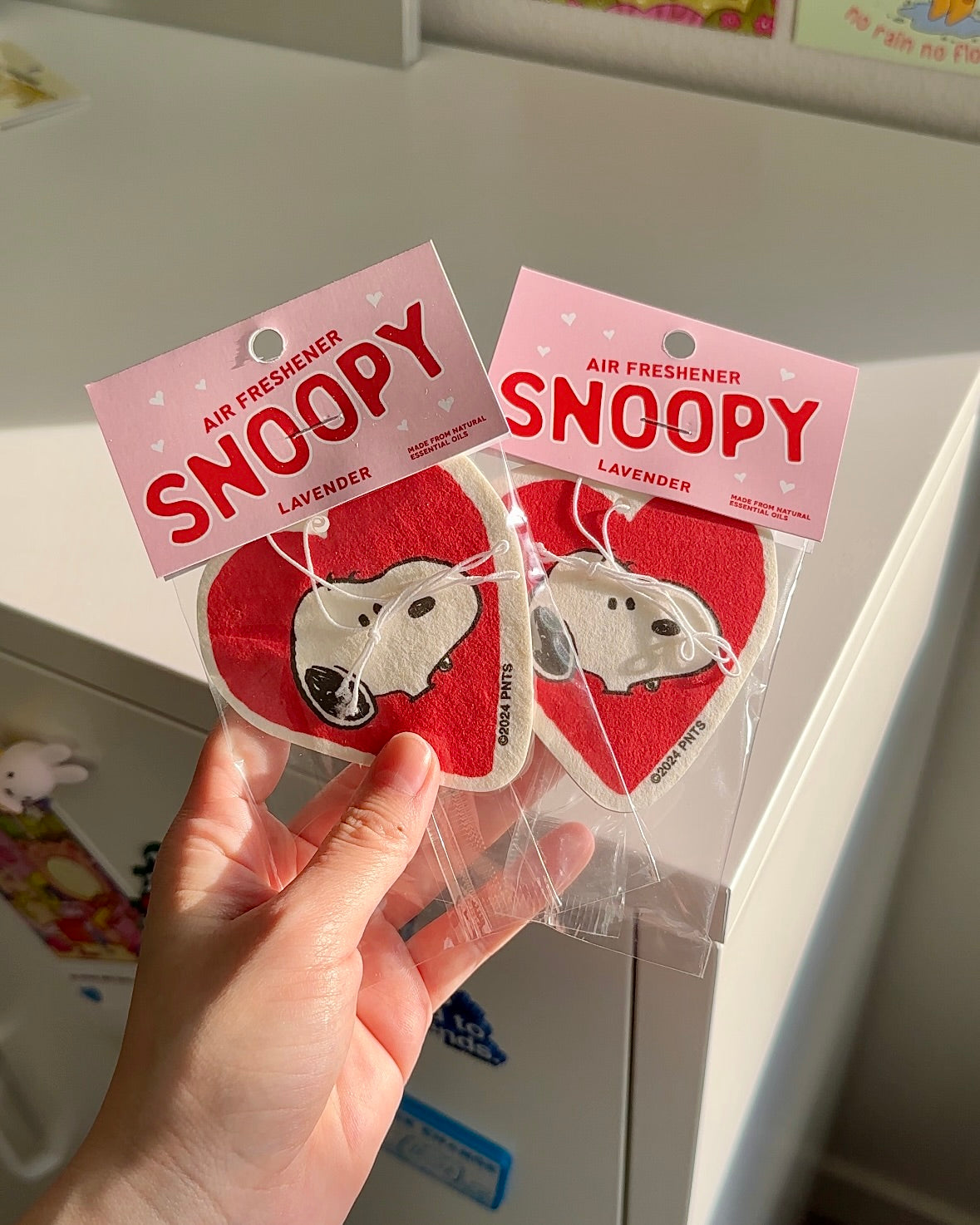 Pair of red heart-shaped Snoopy air fresheners with pink tags, photographed in hand under warm sunlight on a white cabinet.