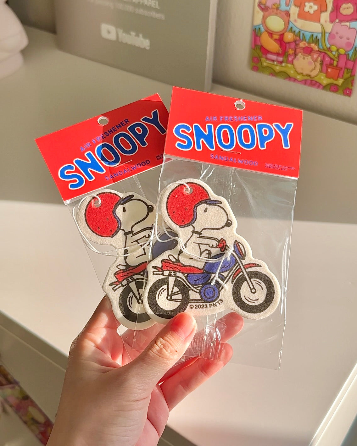 Snoopy motorcycle air freshener in sandalwood scent, shown in packaging. Cute car accessory for Peanuts fans, held in hand with soft lighting.