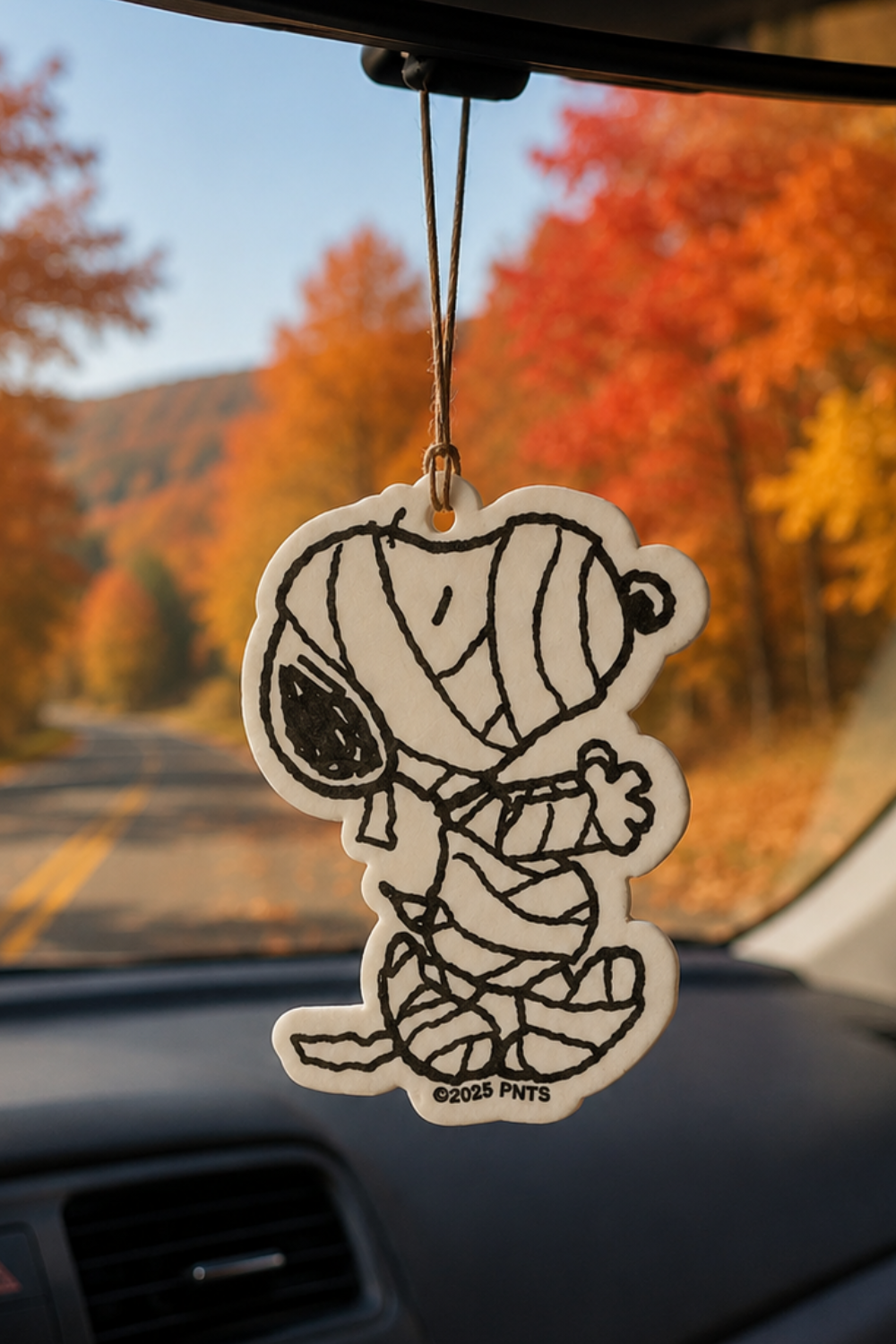 Snoopy Mummy car air freshener with black-and-white mummy costume design, hanging in a car with an autumn mountain road in the background.
