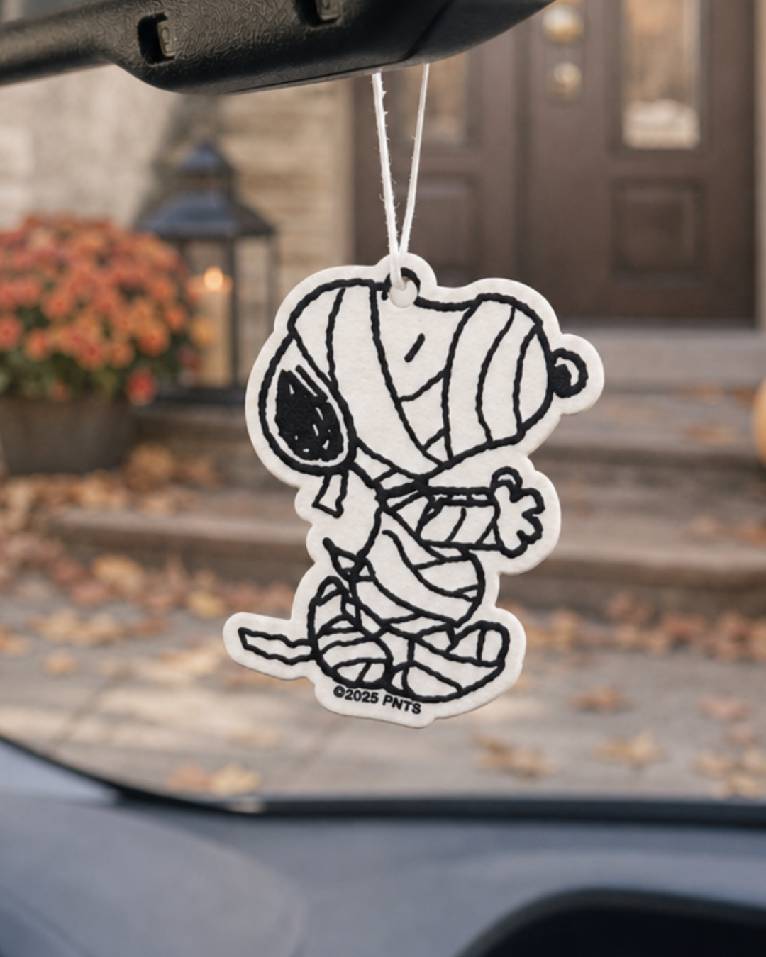 Black and white Snoopy Mummy air freshener hanging from a car mirror in front of a front porch with a lantern and fall flowers.
