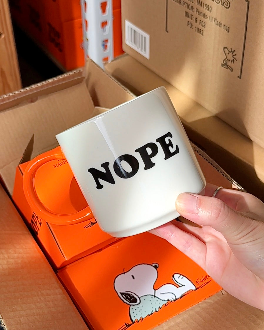 White Snoopy coffee mug with bold “NOPE” text and orange handle, Peanuts ceramic mug held in hand, funny and relatable coffee cup.