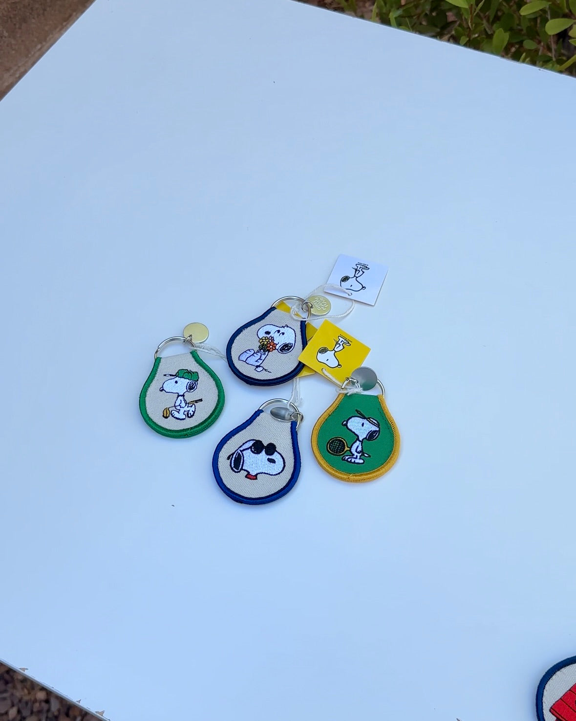 Four Snoopy Peanuts embroidered keychains in green, blue, and yellow trims, featuring tennis Snoopy, sunglasses Snoopy, and Snoopy holding flowers.