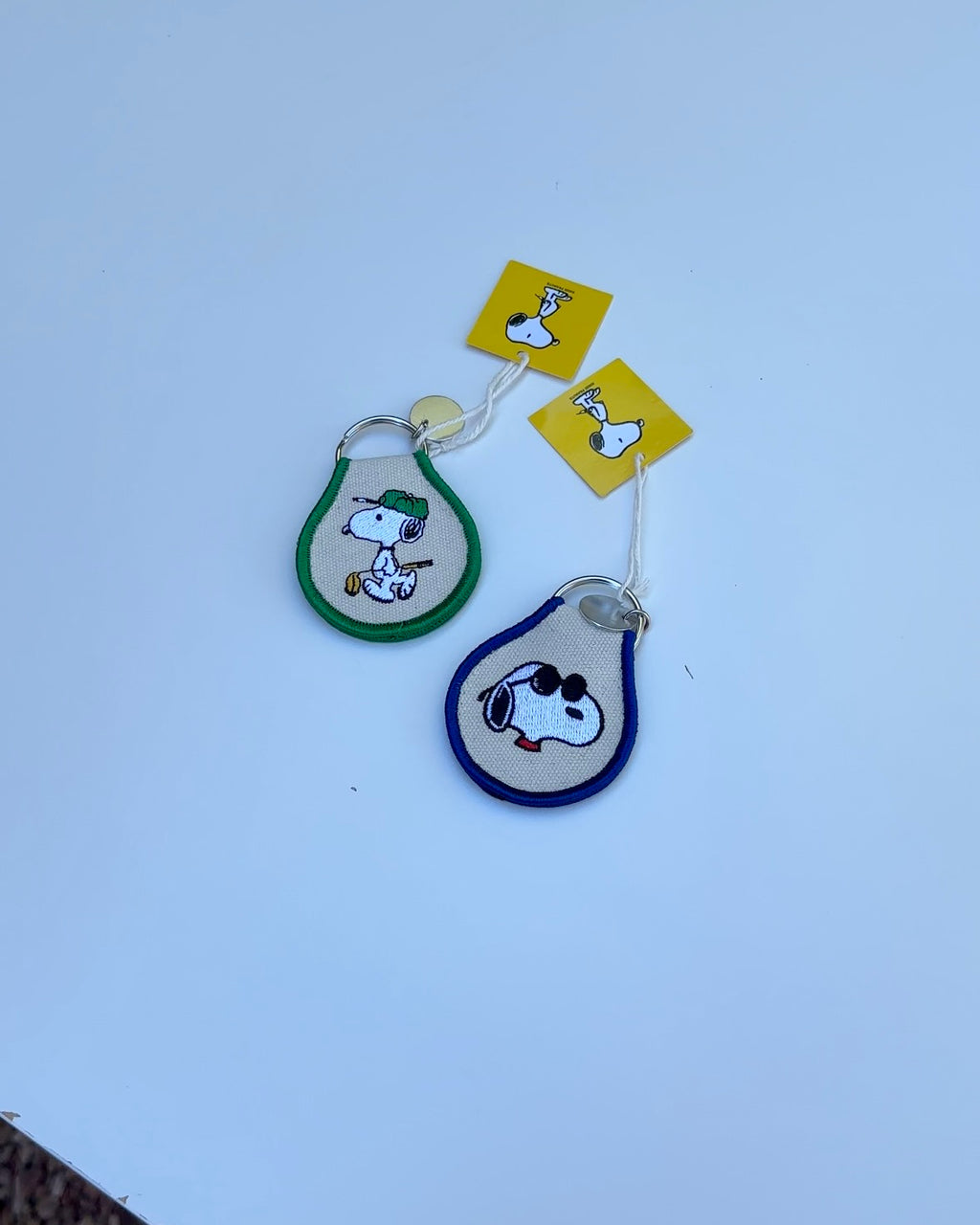 Two Snoopy Peanuts embroidered keychains, one in green with tennis Snoopy and one in blue with sunglasses Snoopy, displayed on a white background.